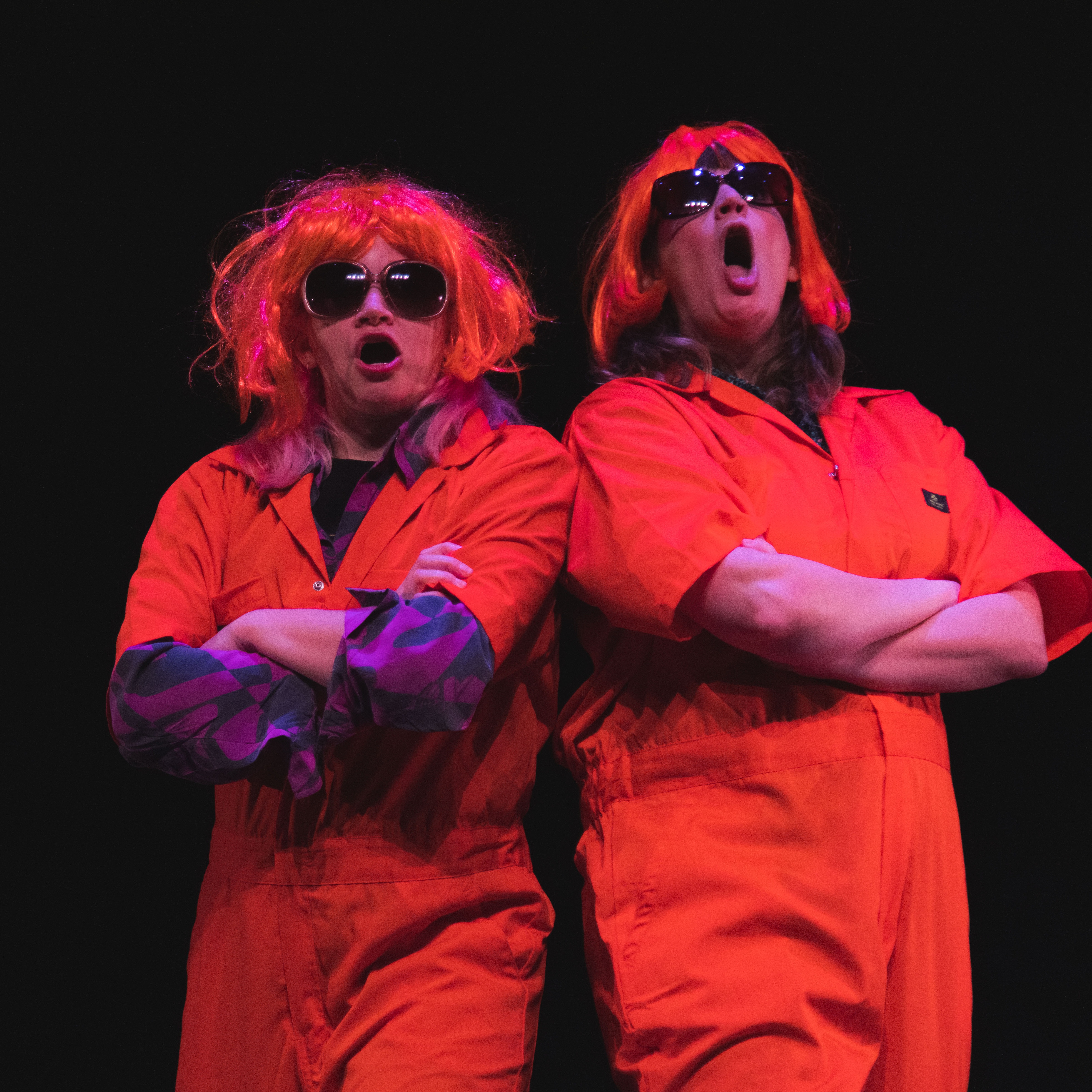 2 white cis women are wearing orange wigs, orange jumpsuits, and sunglasses.  They are yelling and folding their arms.