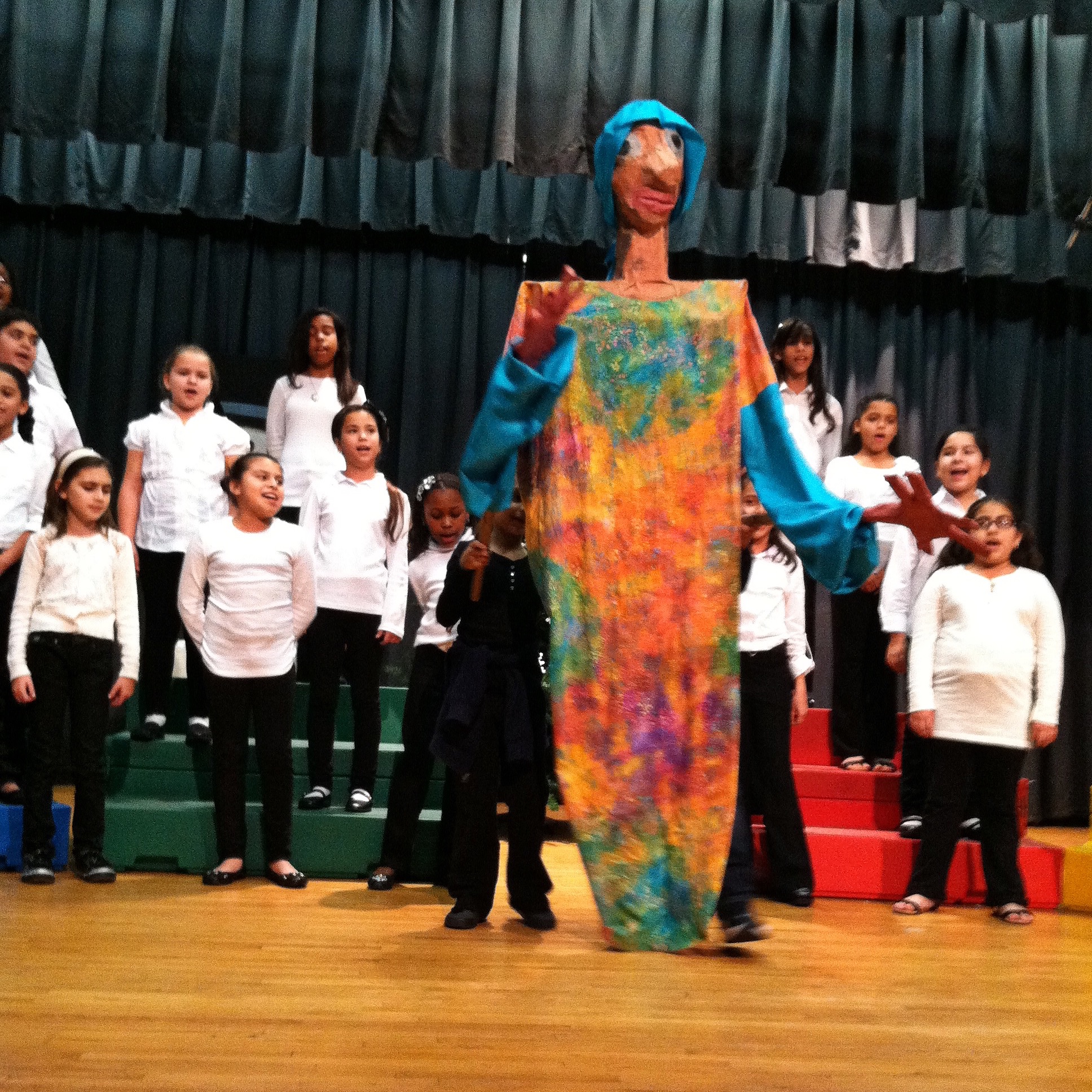 Larger than life puppet made by children in blues and orange colors. A chorus of children singing behind it