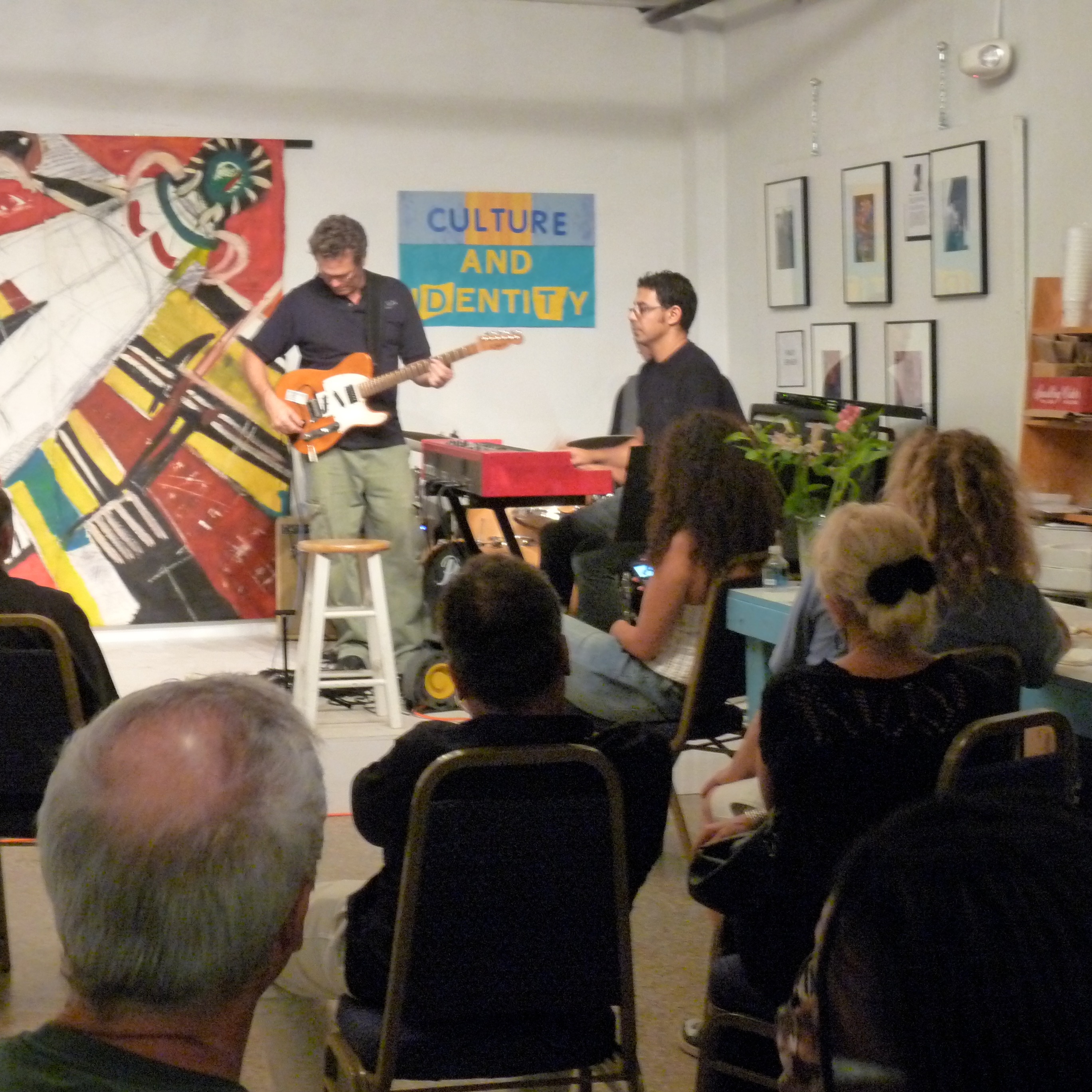 Musicians playing at World and Eye Arts Center with an audience watching and artwork on the walls. The exhibit title is "Culture and Identity".