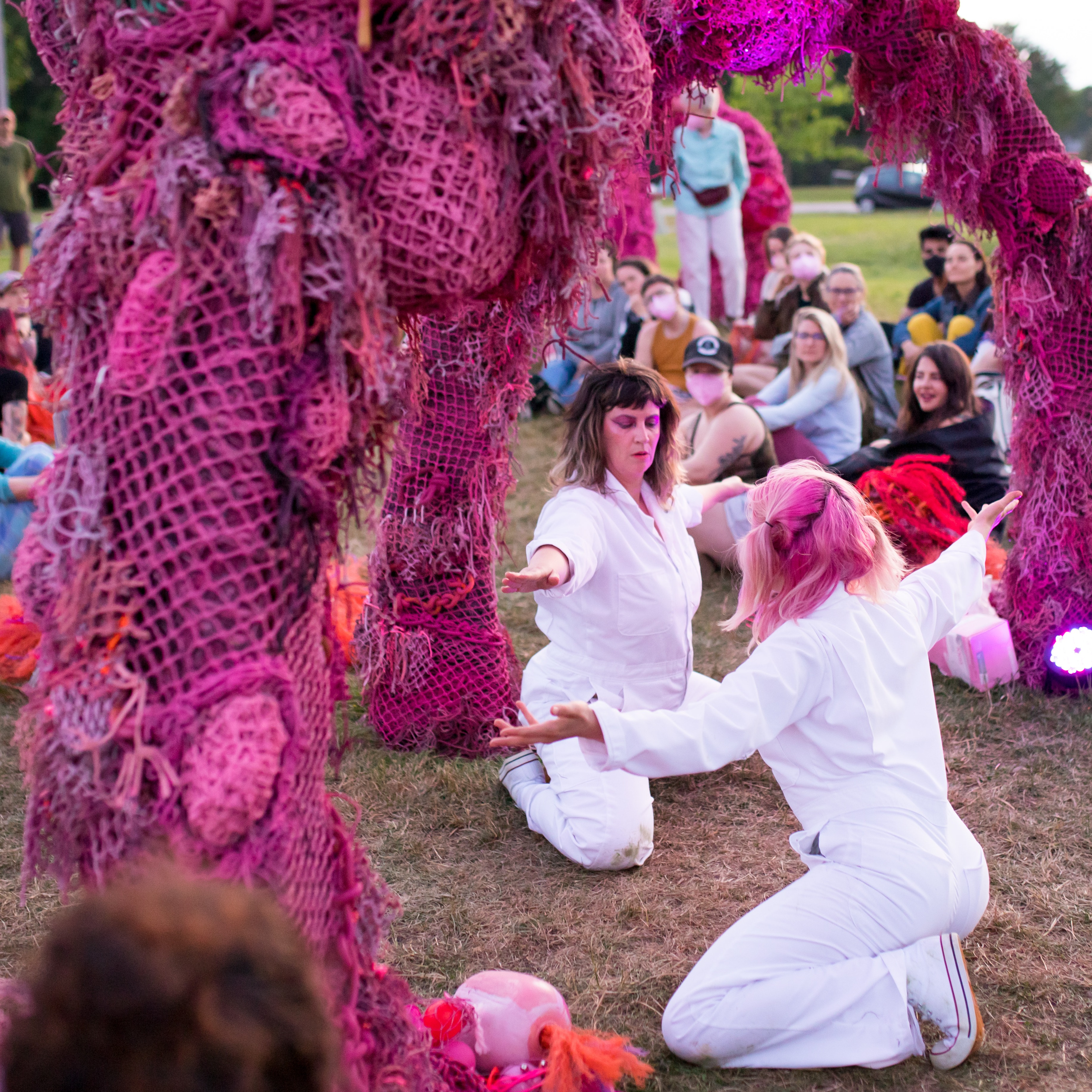 Two white cis women dance under pink rope sculpture with audience watching them