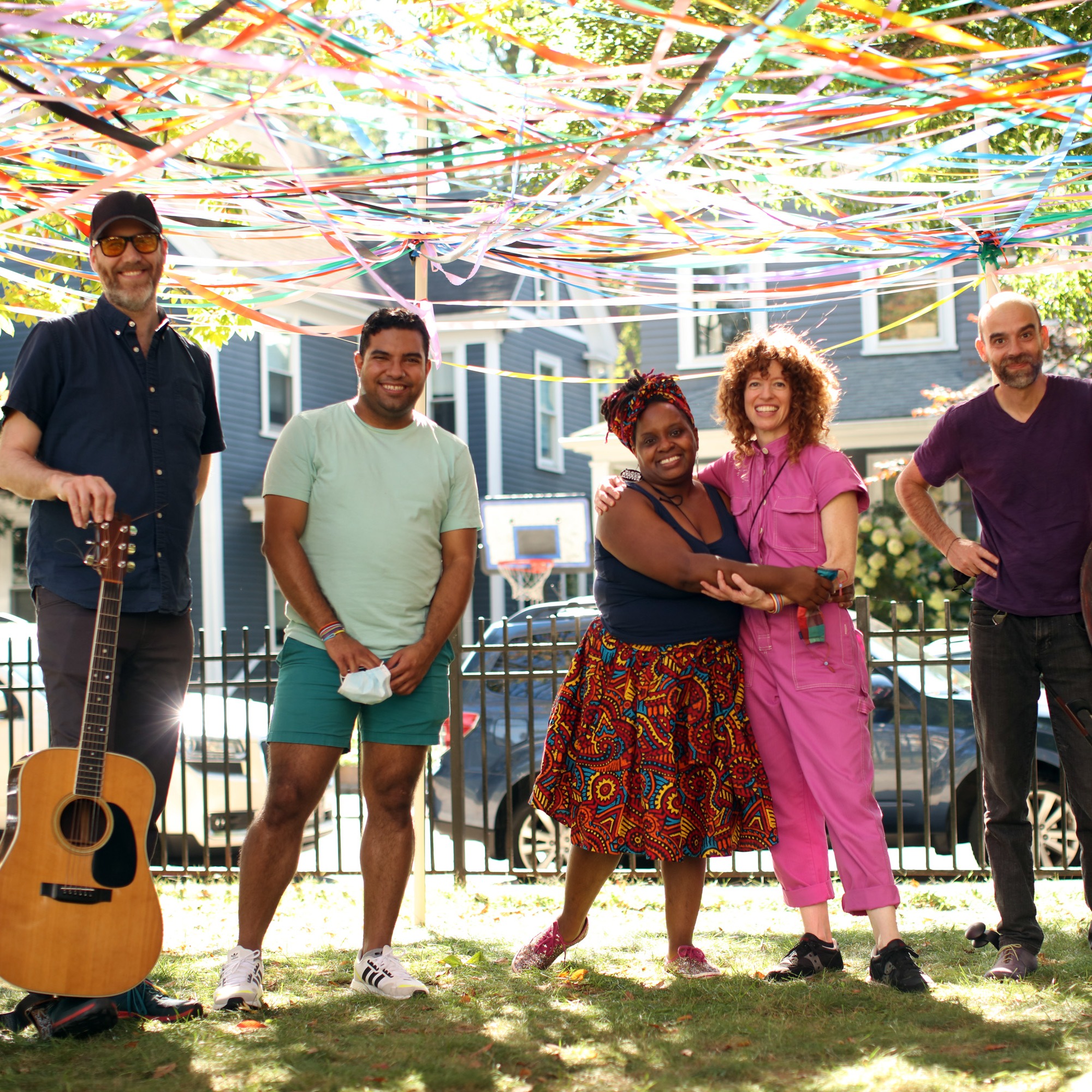 5 performers stand under a canopy of colorful ribbons 