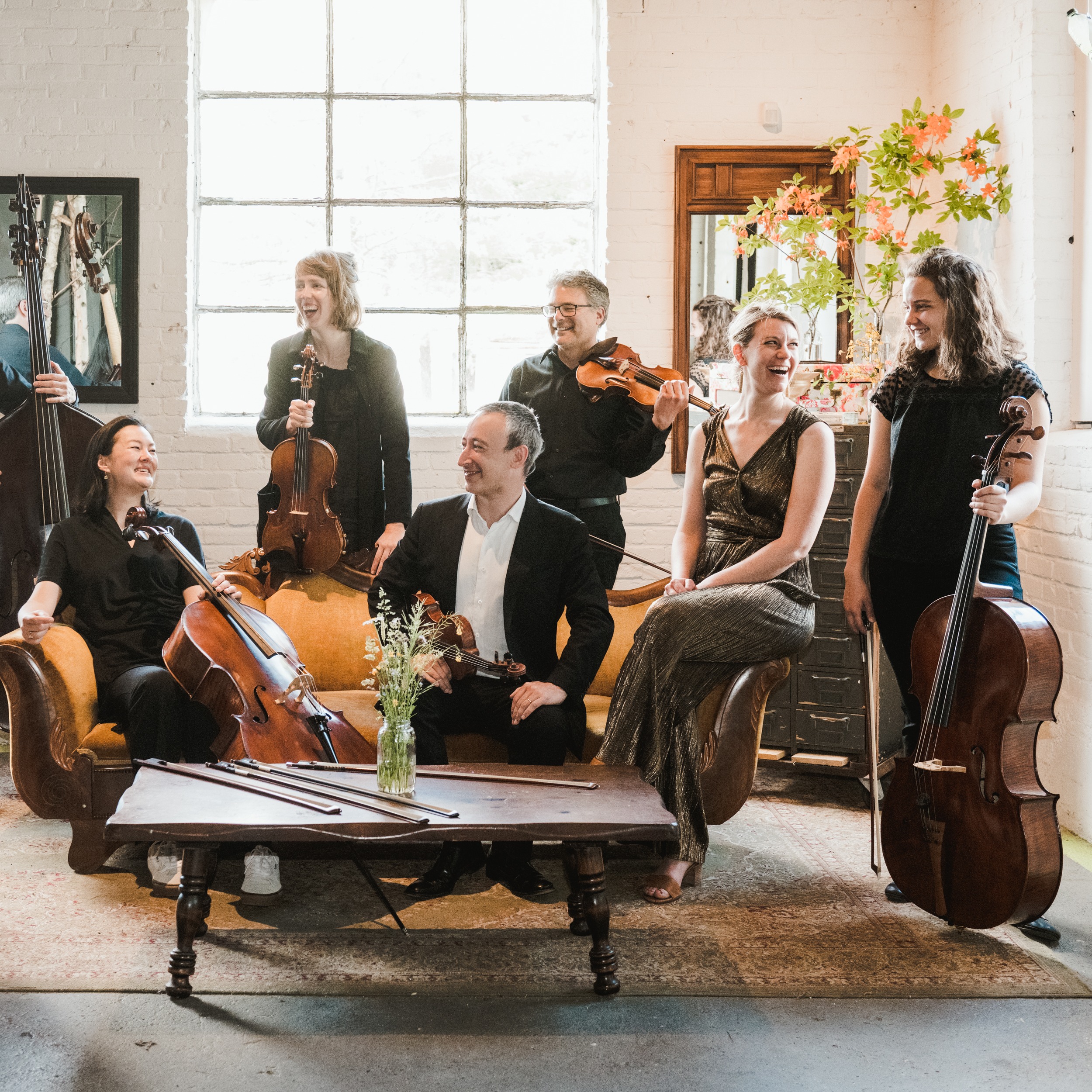 6 individuals with string instruments sitting on and standing around a couch