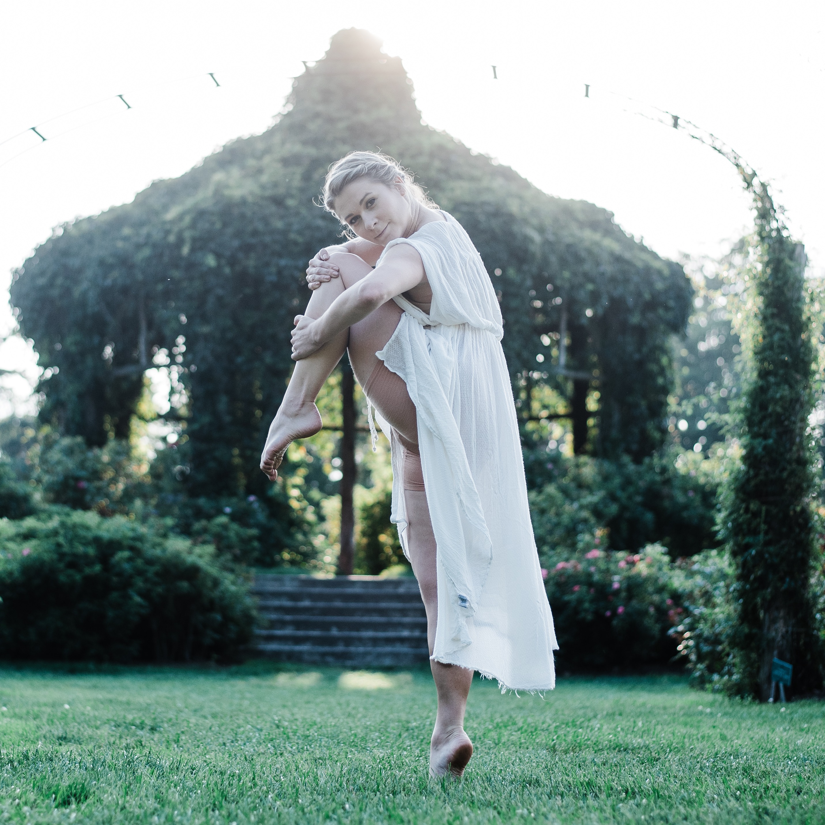 In a lucious garden with a gazebo covered in vines in the back drop, Christina is balancing on one leg with the other tucked into her chest and her focus to the camera. 