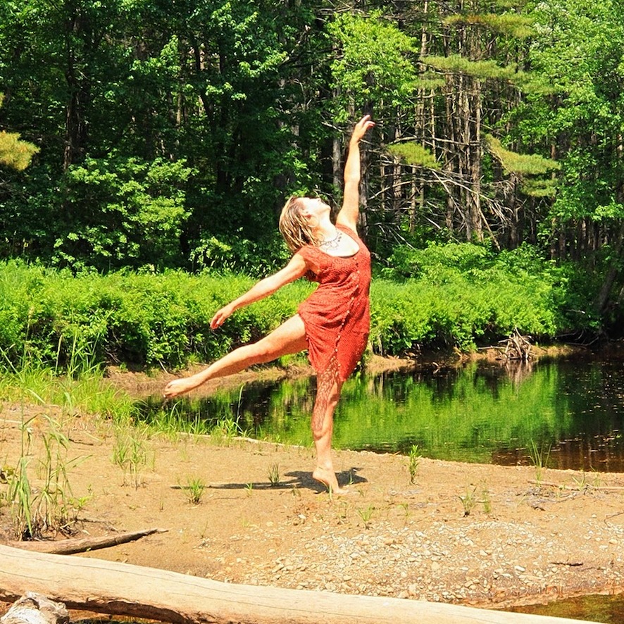 A women with pale skin and damp blonde hair is reaching out legs and arms on a river bend lush with ferns. 
