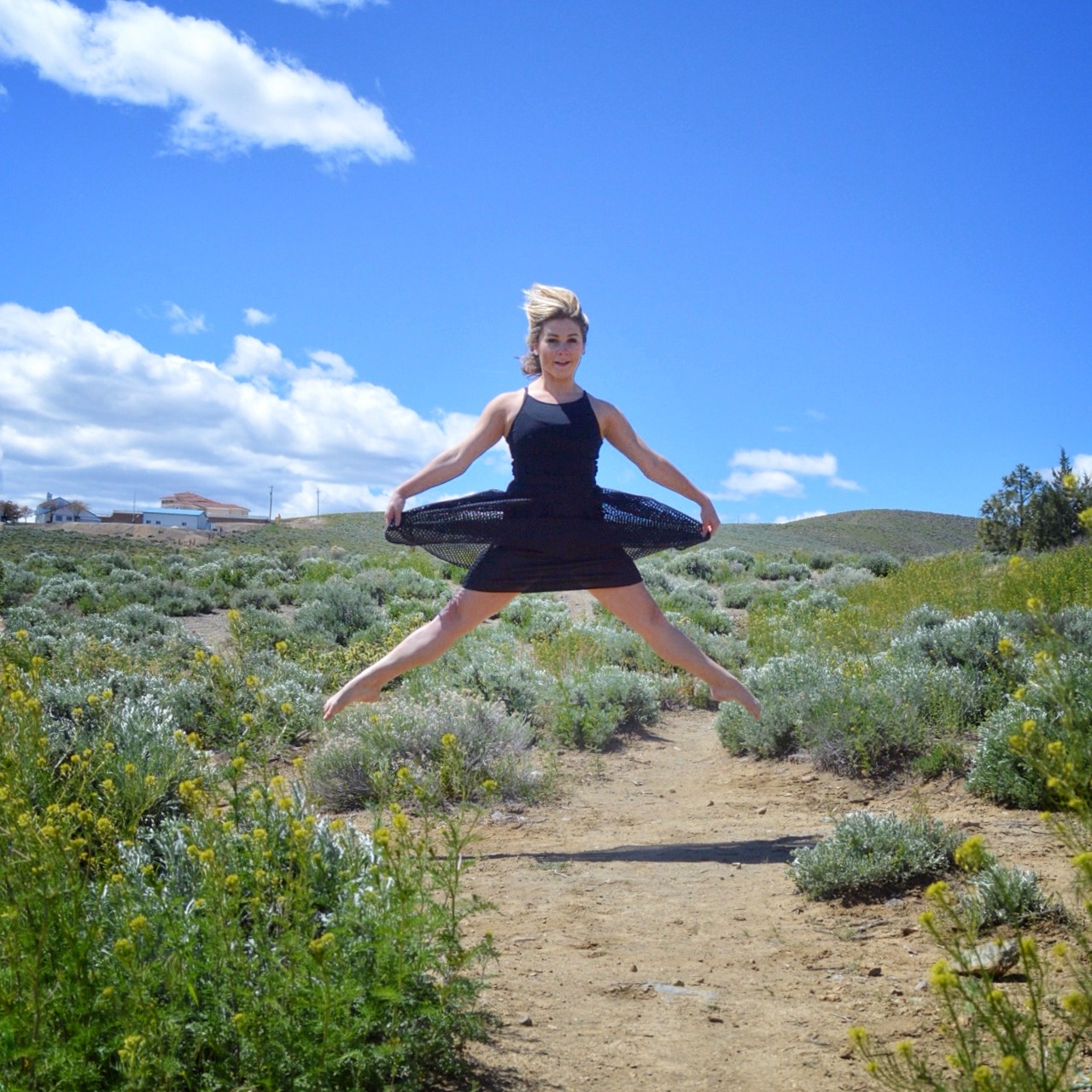 Christina wearing a black dress is floating in the air in a straddle in a high-desert setting.