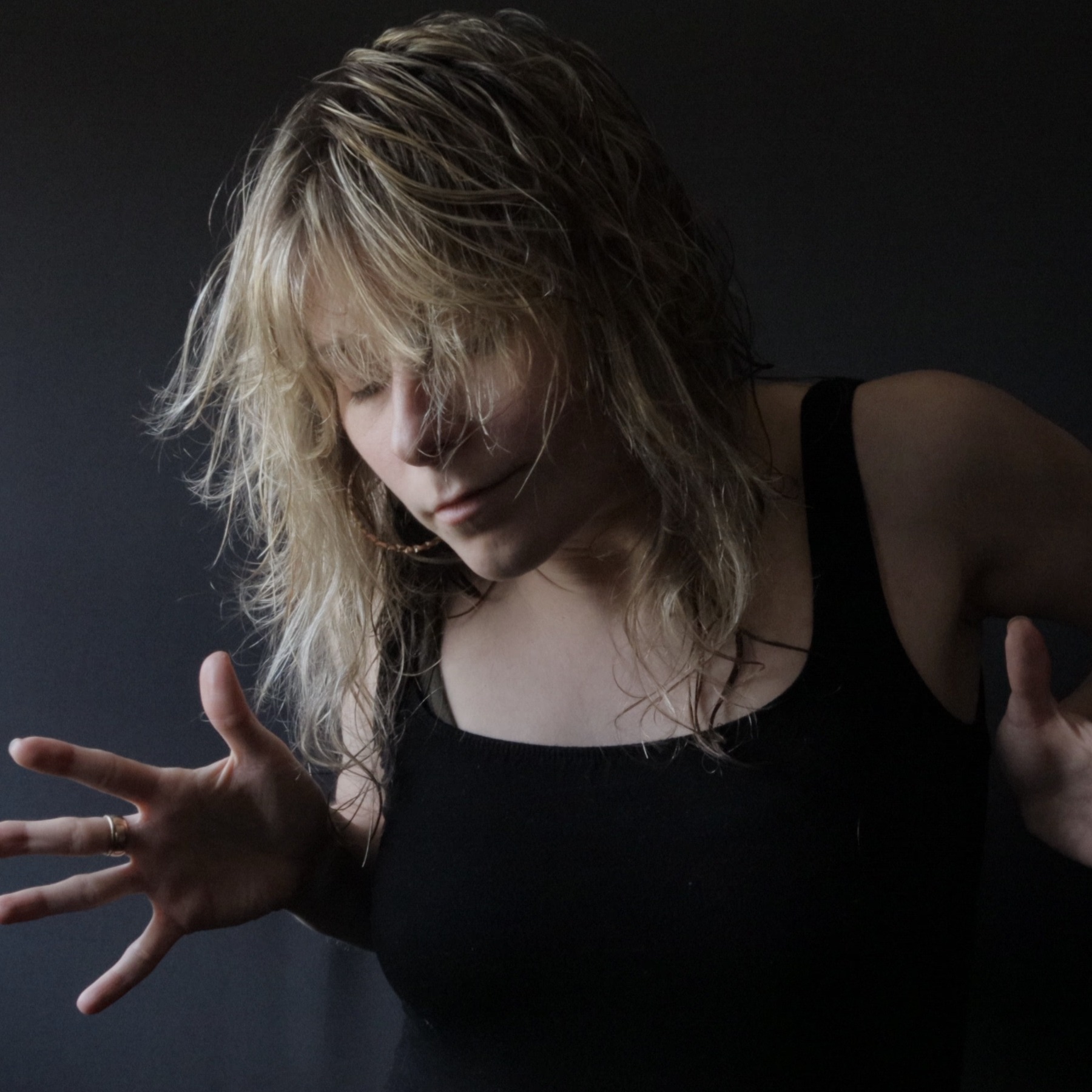 Christina's blonde wavy hair and bangs wisp across her face which looks at peace  yet tension is held in jazz hands energized next to her ribs. The background is black like her tank top. 