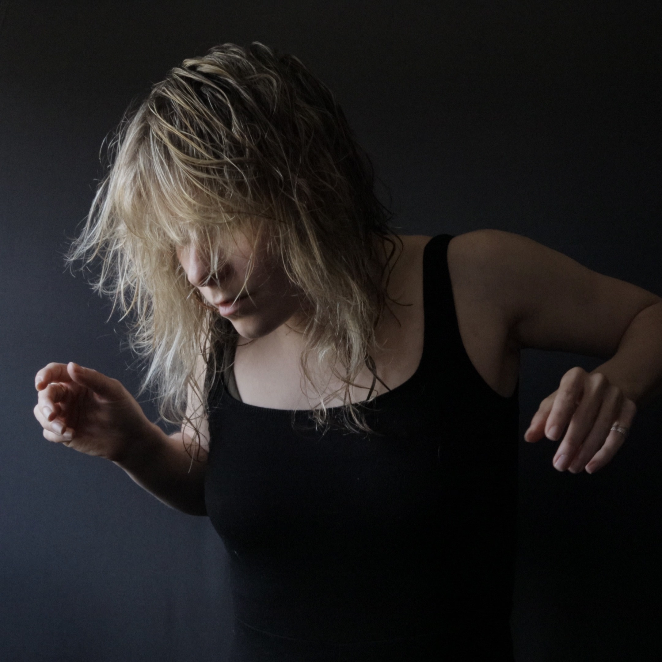 Dressed in black with a black backdrop, Christina's blonde hair covers her face as her arms in motion.