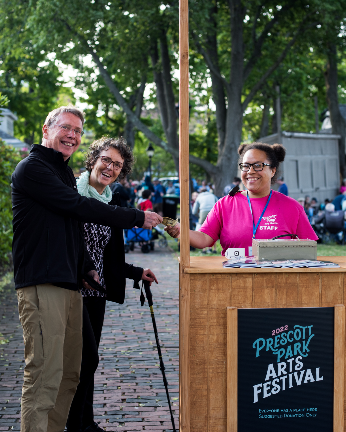 Two people are welcomed at the Arts Festival gate in the park by a gatekeeper wearing a bright pink shirt handing them a season program. All are smiling.  