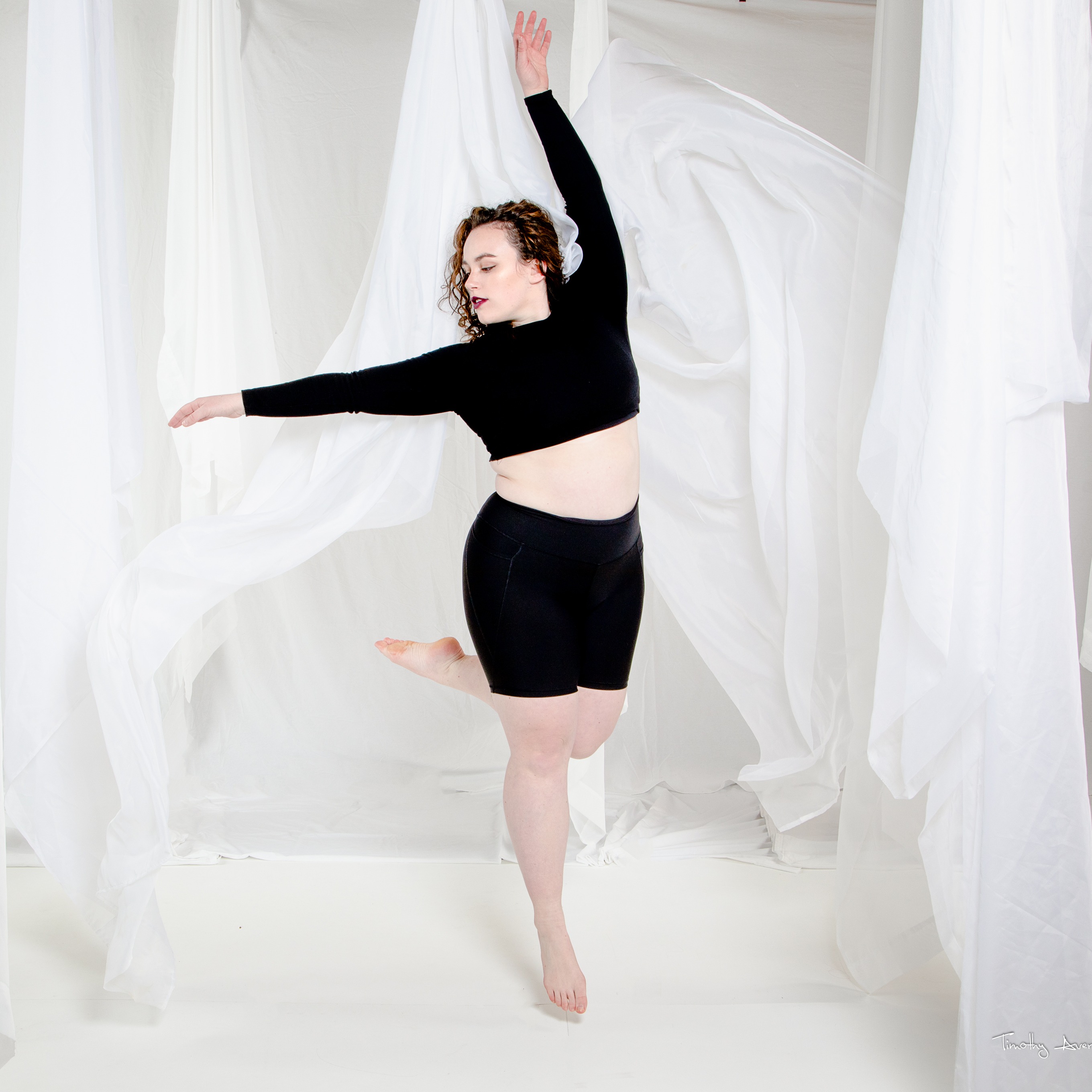 A woman in black shorts and a long sleeve black crop top jumps in white fabric with one leg swinging behind her and one arm over her head.