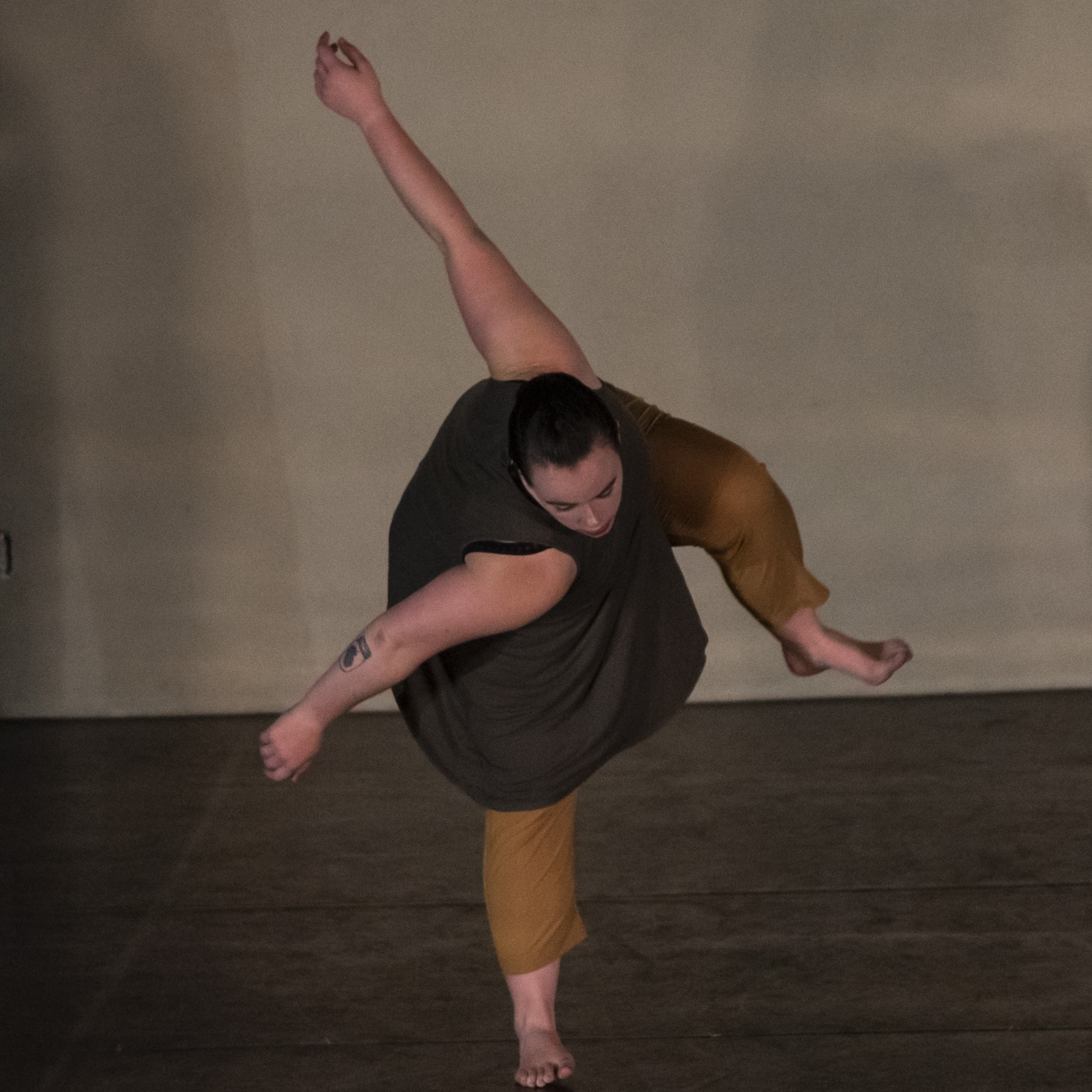 A woman in a green top and yellow pants lefts her left leg and arm while bent over to look at the ground.