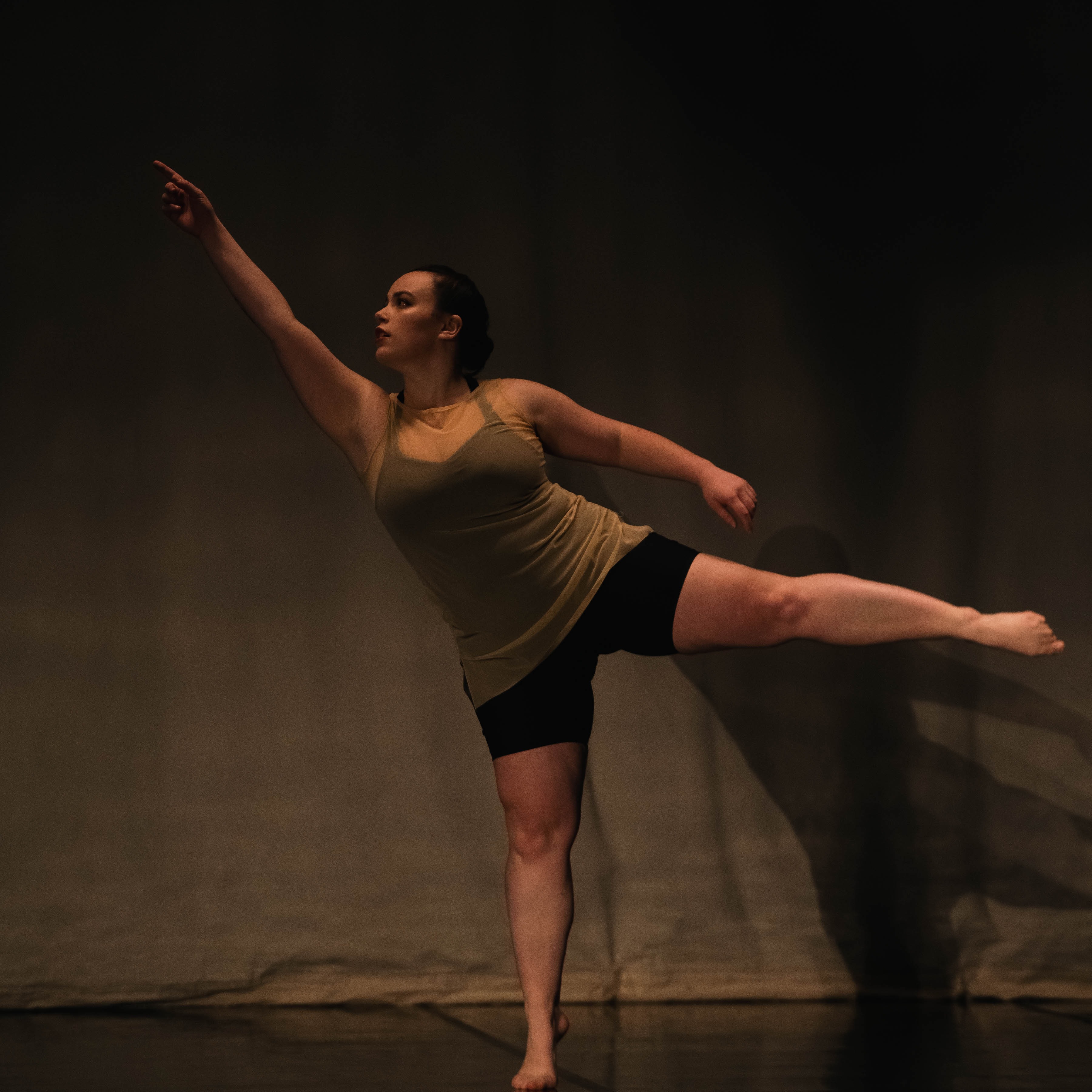 A woman in a sheer nude dress over black shorts reaches her right arm high while her left leg extends to the side.