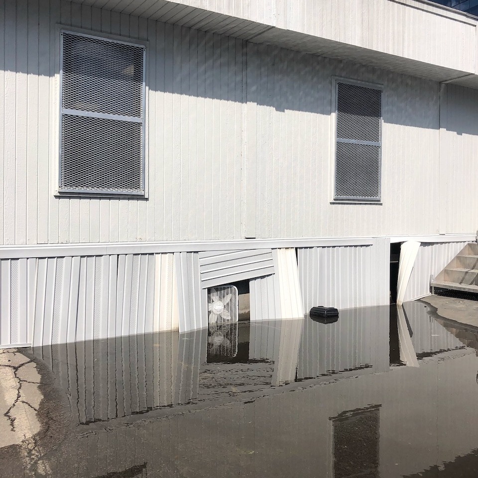 An image of a building that is partway under water, and that water creates a mirror image of what is above it. There is a box fan partly submerged and mirrored in the water.