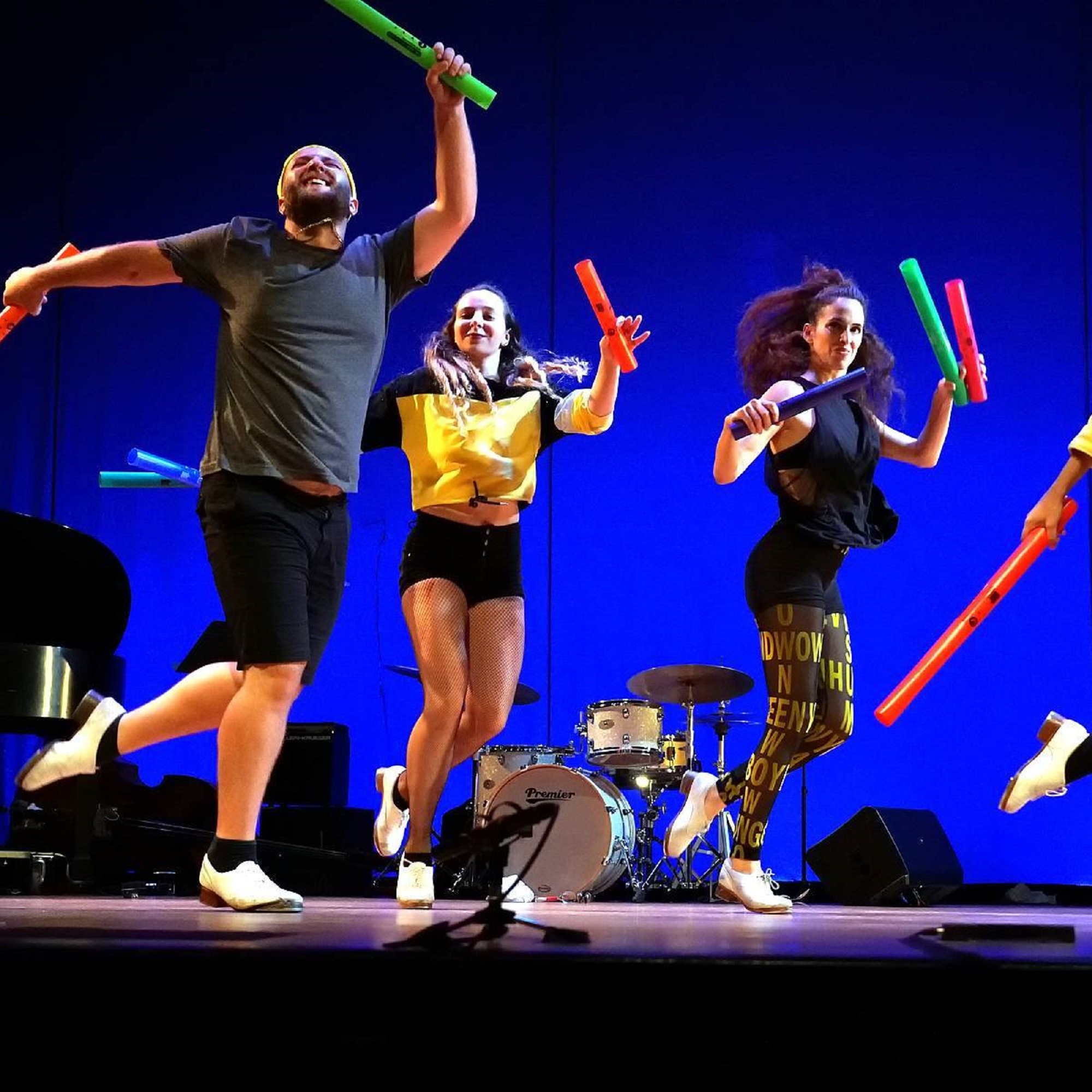 Four dancers dressed in black, yellow and grey, with tap dance shoes, on stage with a blue background. The dancers are holding colorful plastic pipes.