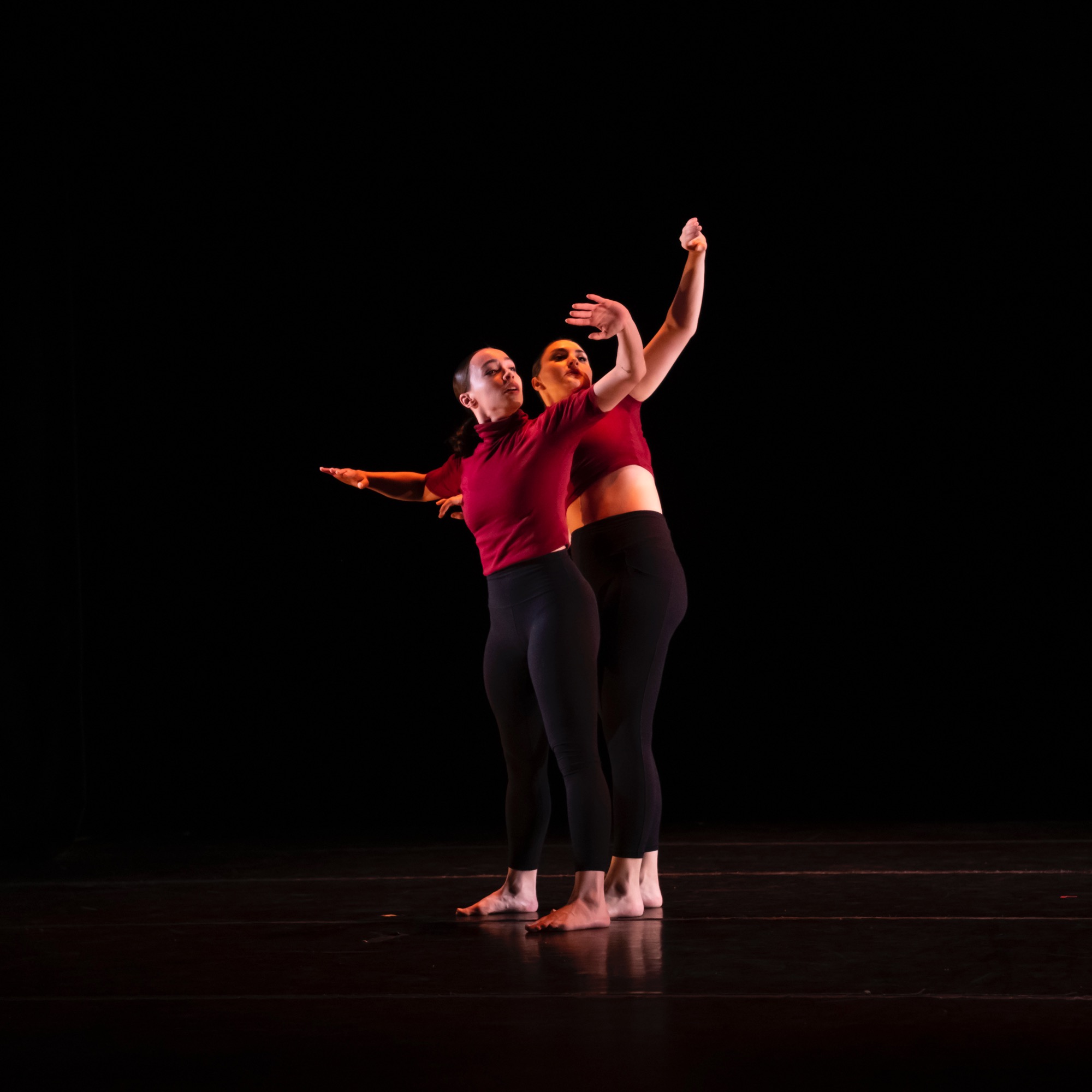 Two dancers stand one behind the other with arms extended upward 