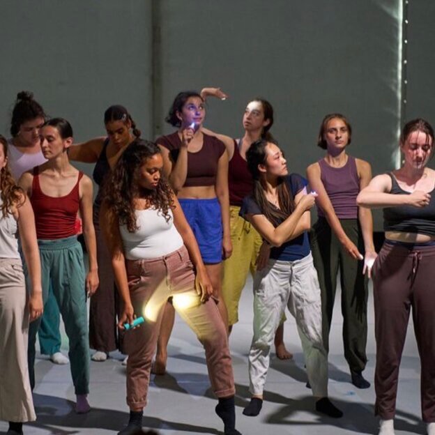10 dancers are dressed in solid colored, jewel toned tops and bottoms. The dancers are pointing a white, green, blue or green light at their bodies. They look like they have discovered something i’m their light. 