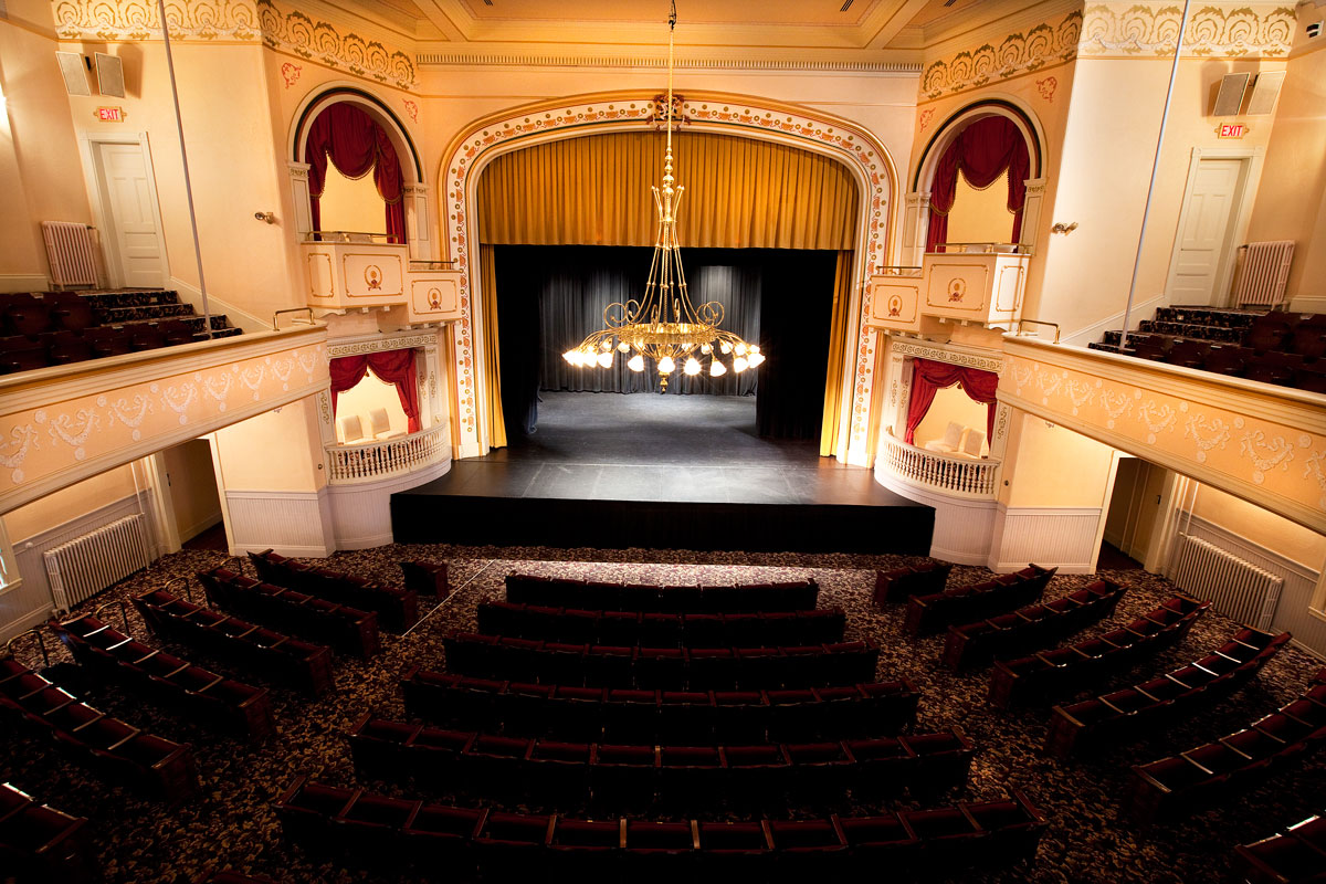 A birds eye view of the Camden Opera House, on the lower half six rows of black chairs, in the middle a black stage with a chandelier above. There's a gold colored curtain above the stage.
