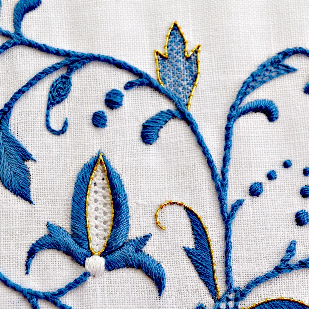 Closeup of white linen embroidered with a trailing design in blue, white, and accented with gold. Several embroidery techniques such as pulled-thread work, couching, and padded satin stitch are shown.