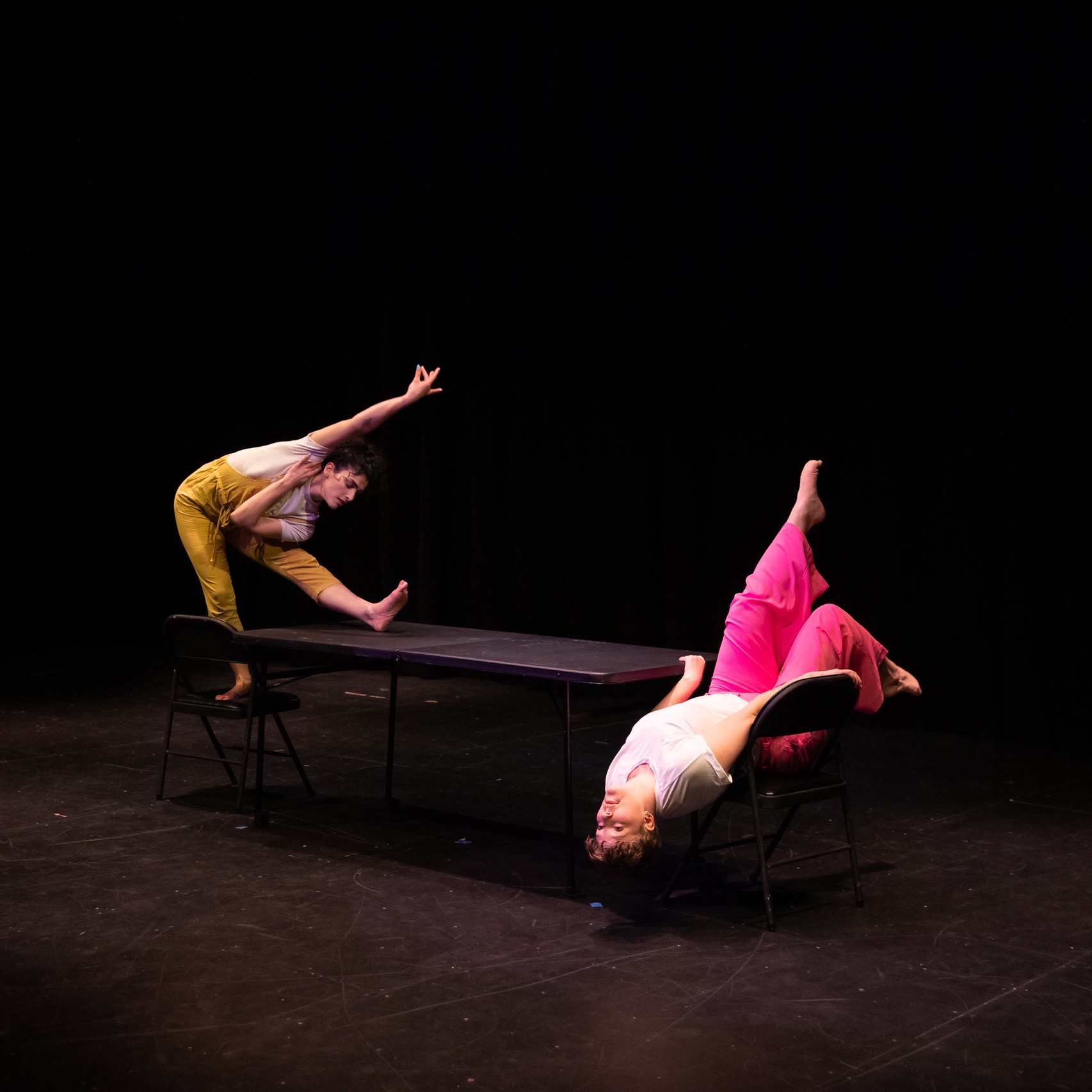 Two people on opposite sides of a table. One standing on a chair bent over a foot on the table. The other laying back on a chair with her feet in the air
