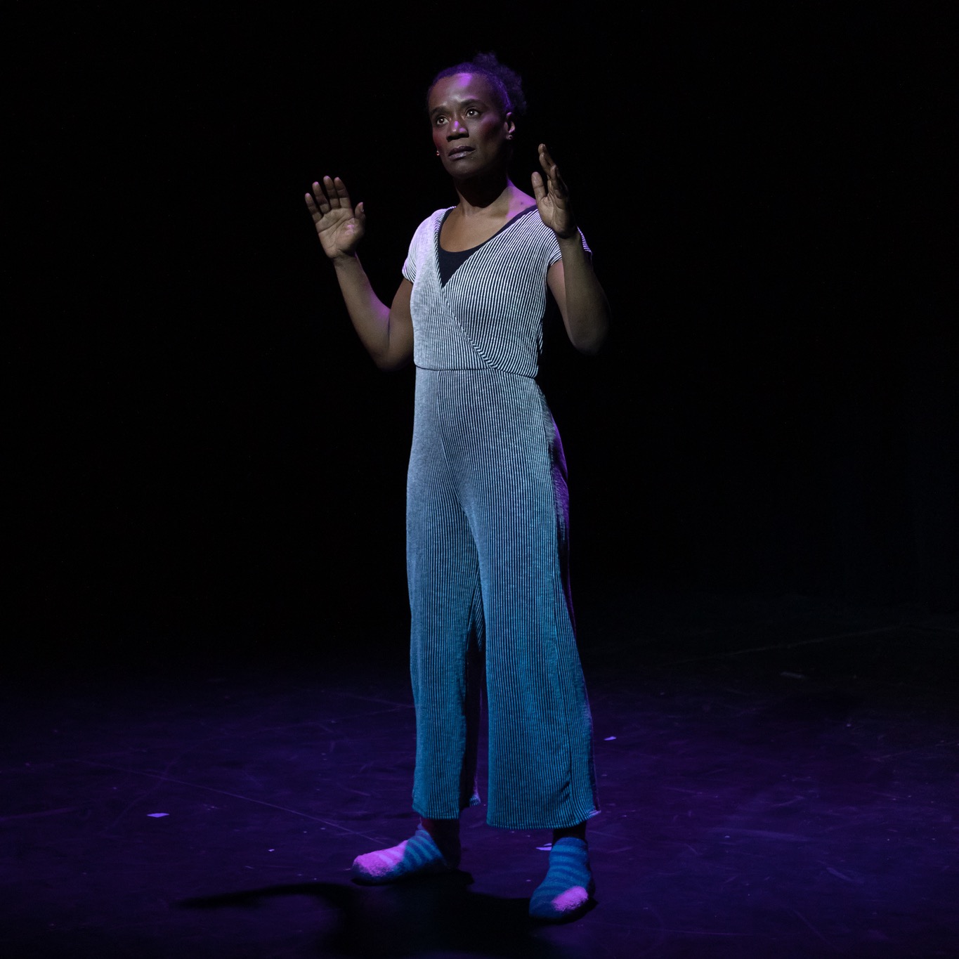 Black woman in a silvery blue jumpsuit standing with her hands raised to her shoulders
