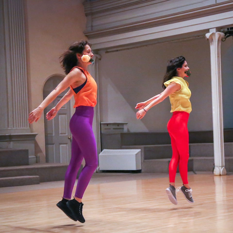 Two dancers in unison, mid chasse, so both feet are off the floor, their arms straight and low behind them. One dancer in bright orange top and purple shiny leggings while the other is in bright yellow top and shiny red pants. 