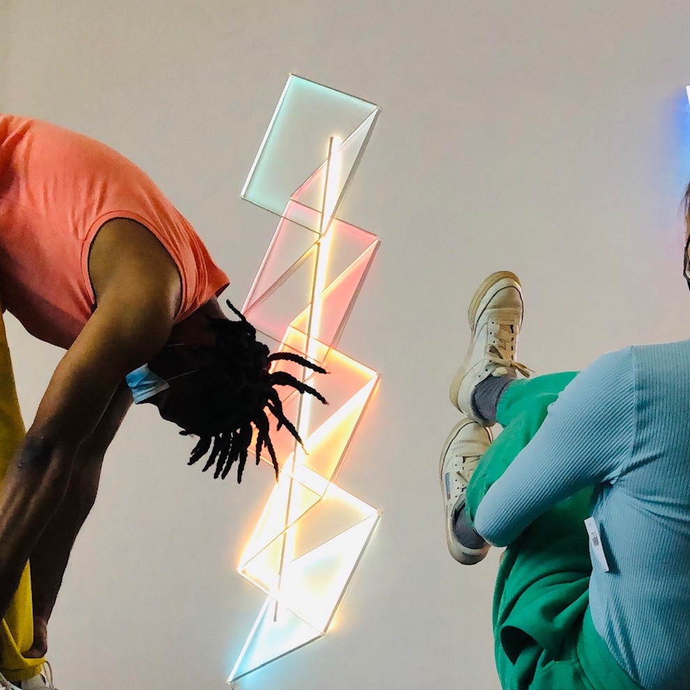 Two bodies are framing and in the forefront to a vertical light sculpture of triangles.  