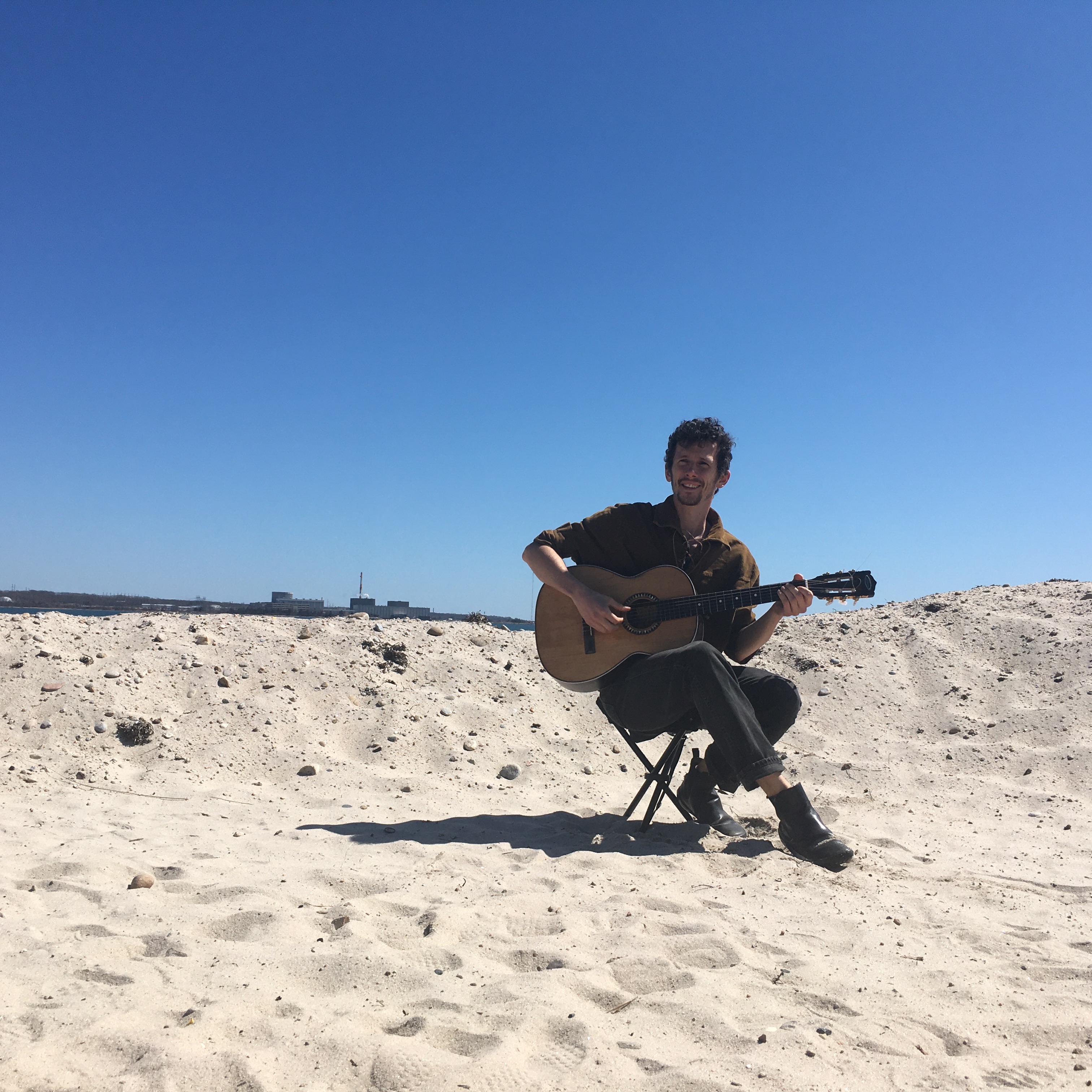 The artist, playing guitar on a small stool on the sand, with a bright blue sky