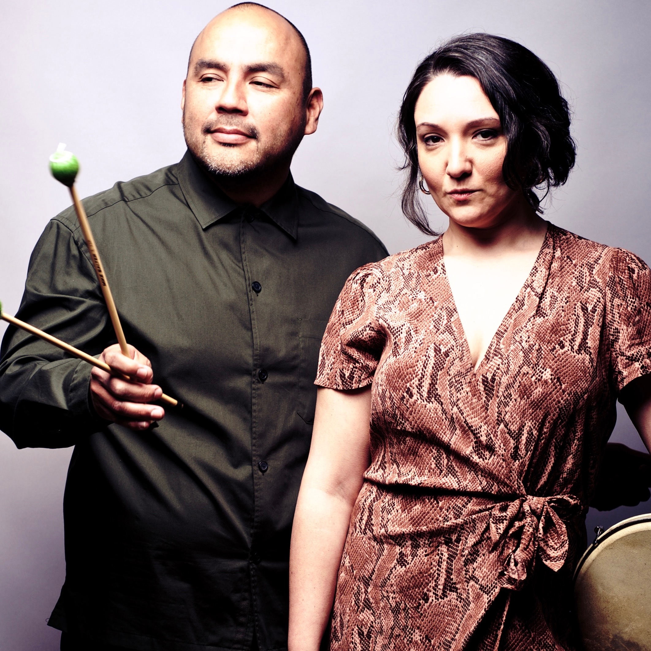 The Bahué Duo, Aliana de la Guardia in the foreground with a pink snake skin print dress, and Ariel Campos in the background with a green button up shirt holding marimba mallets.