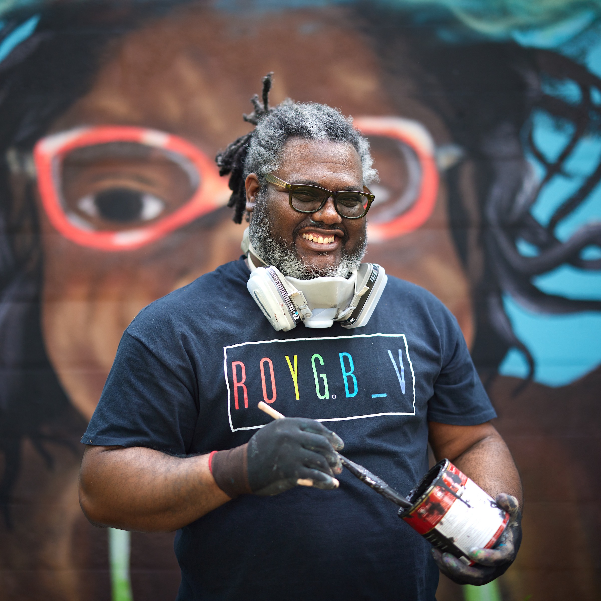 Andre Rochester standing in front of his mural holding a brush and can of paint.
