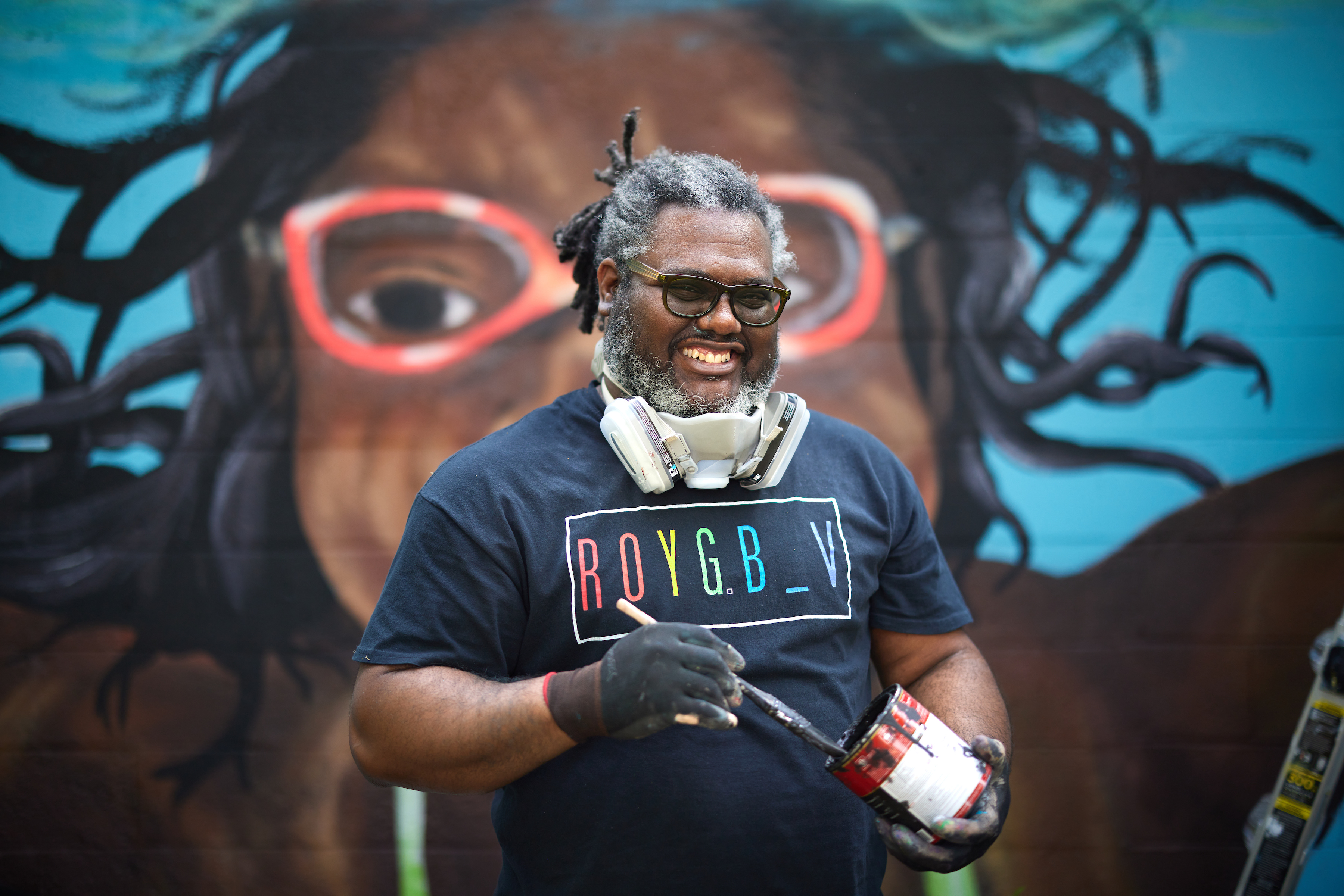 Andre Rochester standing in front of his mural holding a brush and can of paint.