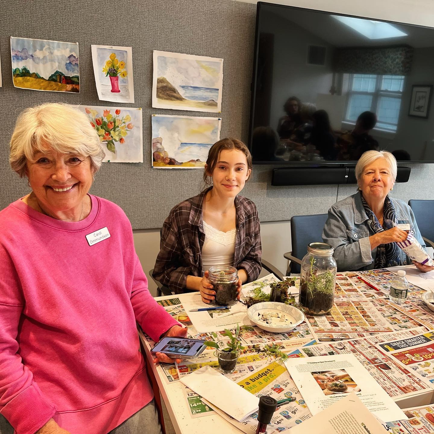 Engaged in crafting terrariums, three individuals share smiles. Among them are two residents from Riverwoods and an Arts In Reach teenager, all collaborating with enthusiasm.