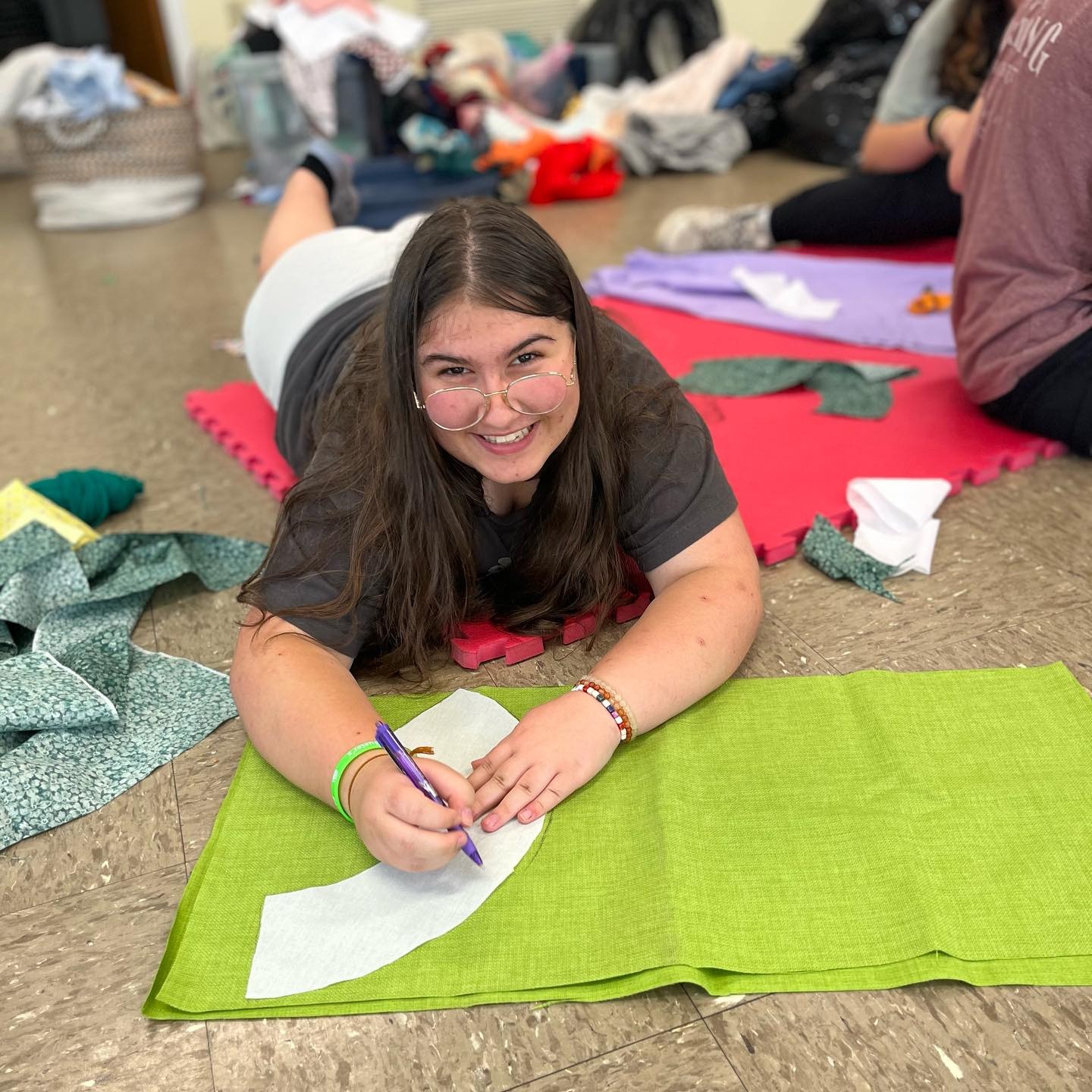 Graced with a smile, a glasses-wearing teenager flaunting long brown hair meticulously traces a portion of a bucket hat pattern onto vibrant lime green fabric.