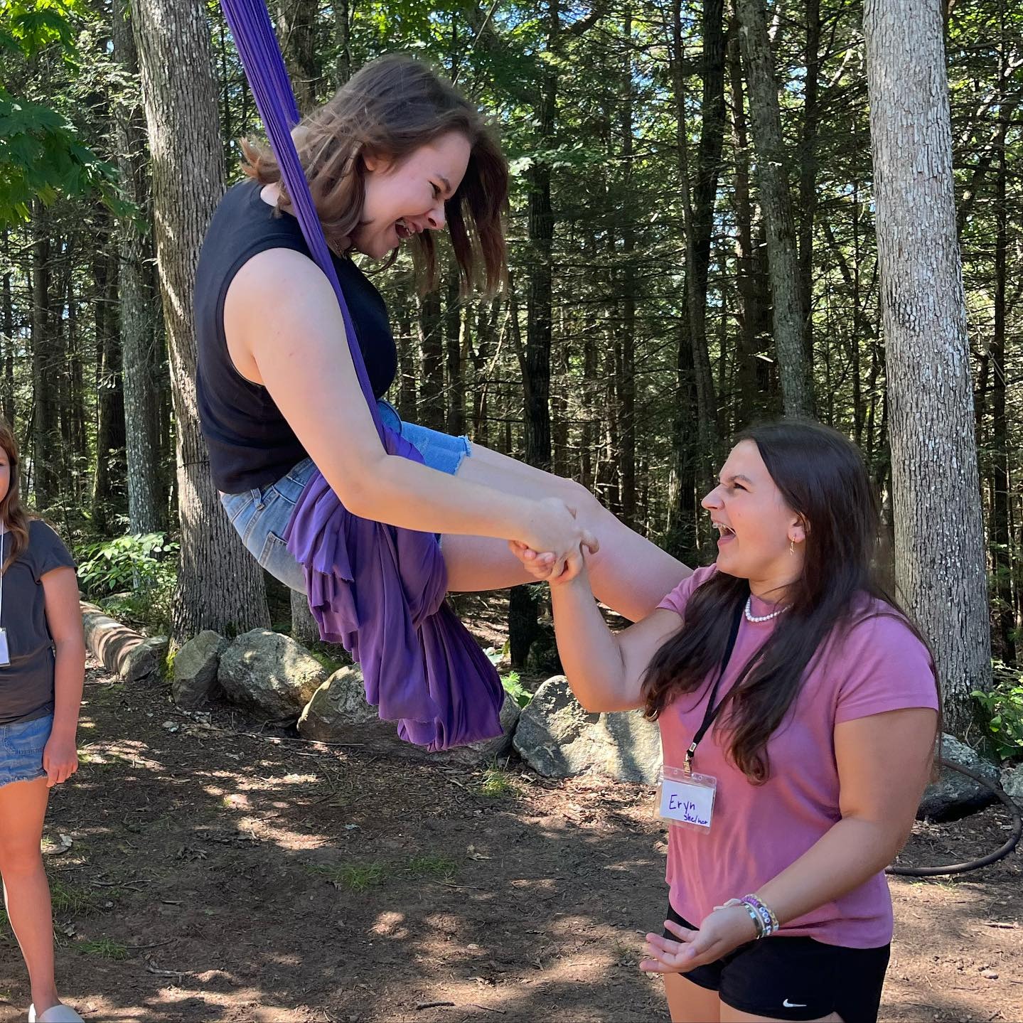 Two smiling teenagers grasp each other's hands joyously. One, with medium-length hair, is suspended on a purple aerial silk in midair, gazing downward at the other teen. The second teenager boasts long brunette hair and wears a pink shirt.