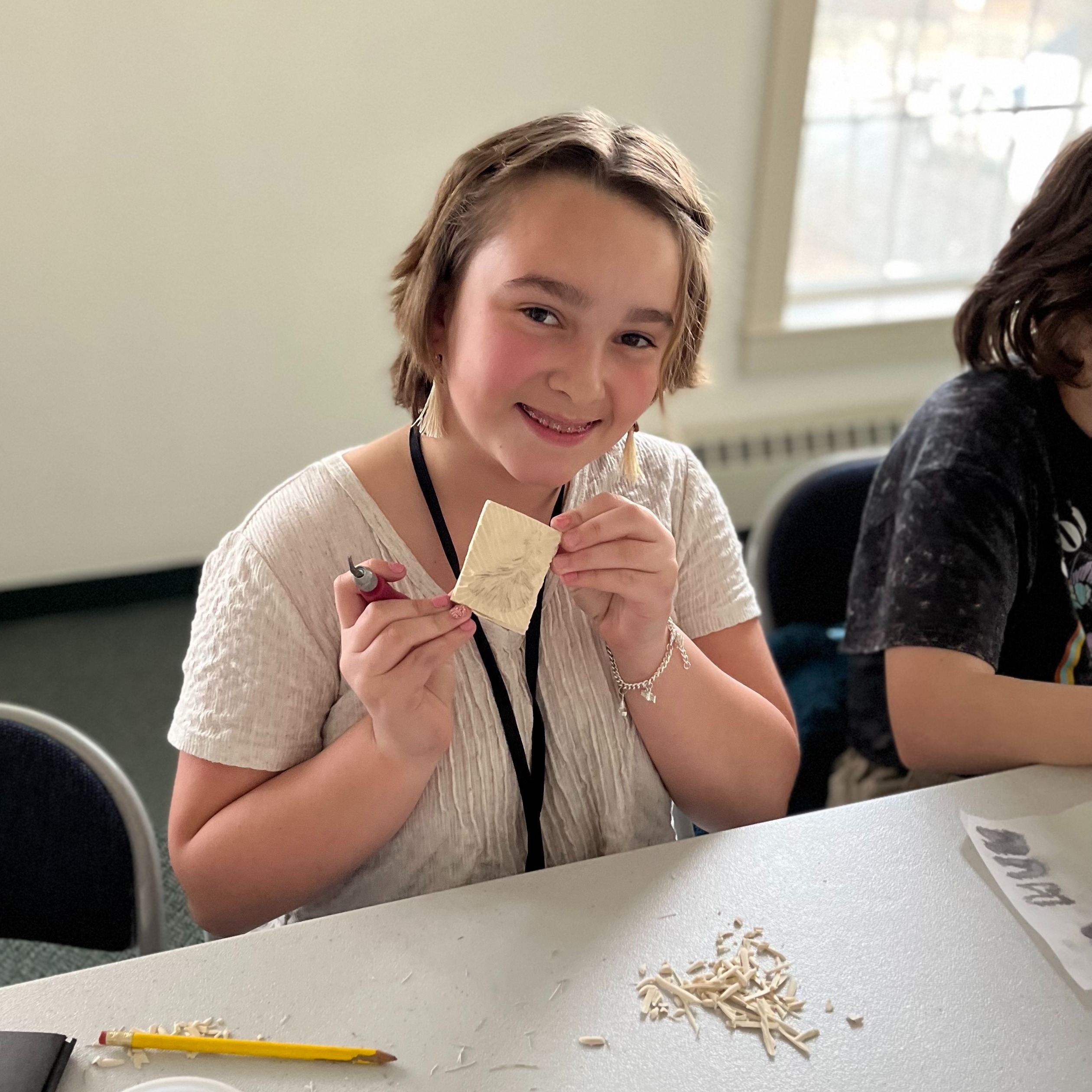 Clad in a white shirt, a teenager with short blonde hair wears a smile as they grasp a partially carved linoleum block. This block is destined to produce patches once printed.