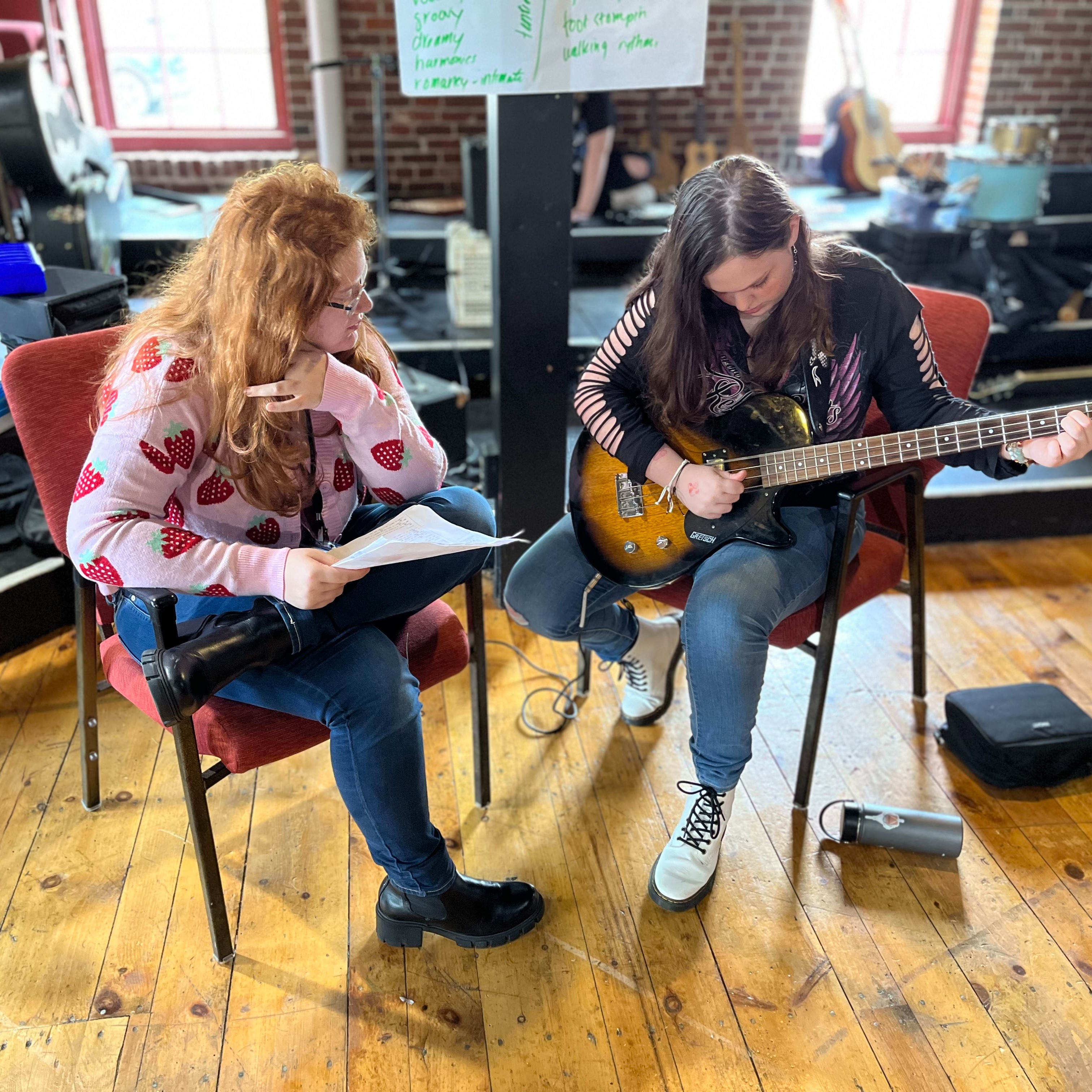 A teenager with flowing brunette hair strums a bass guitar in shades of black and brown, while a second teen sporting long red hair and a strawberry-themed cardigan gazes upon the instrument with interest.