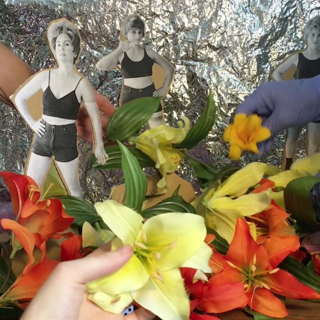 Colorful flowers fill a foreground, and three black and white cutout images of white women in cut offs and crop tops stand against a tinfoil backdrop. Hands reach into the frame, one wearing lavender gloves, adjusting the flowers and the cutouts. 