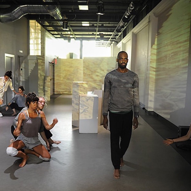 A gallery space has yellow, rippled video projections covering its walls. 6 dancers are in the space—two pairs of dancers are touching, one pair crouched on the ground. One dancer stands looking straight out ahead towards the camera. 