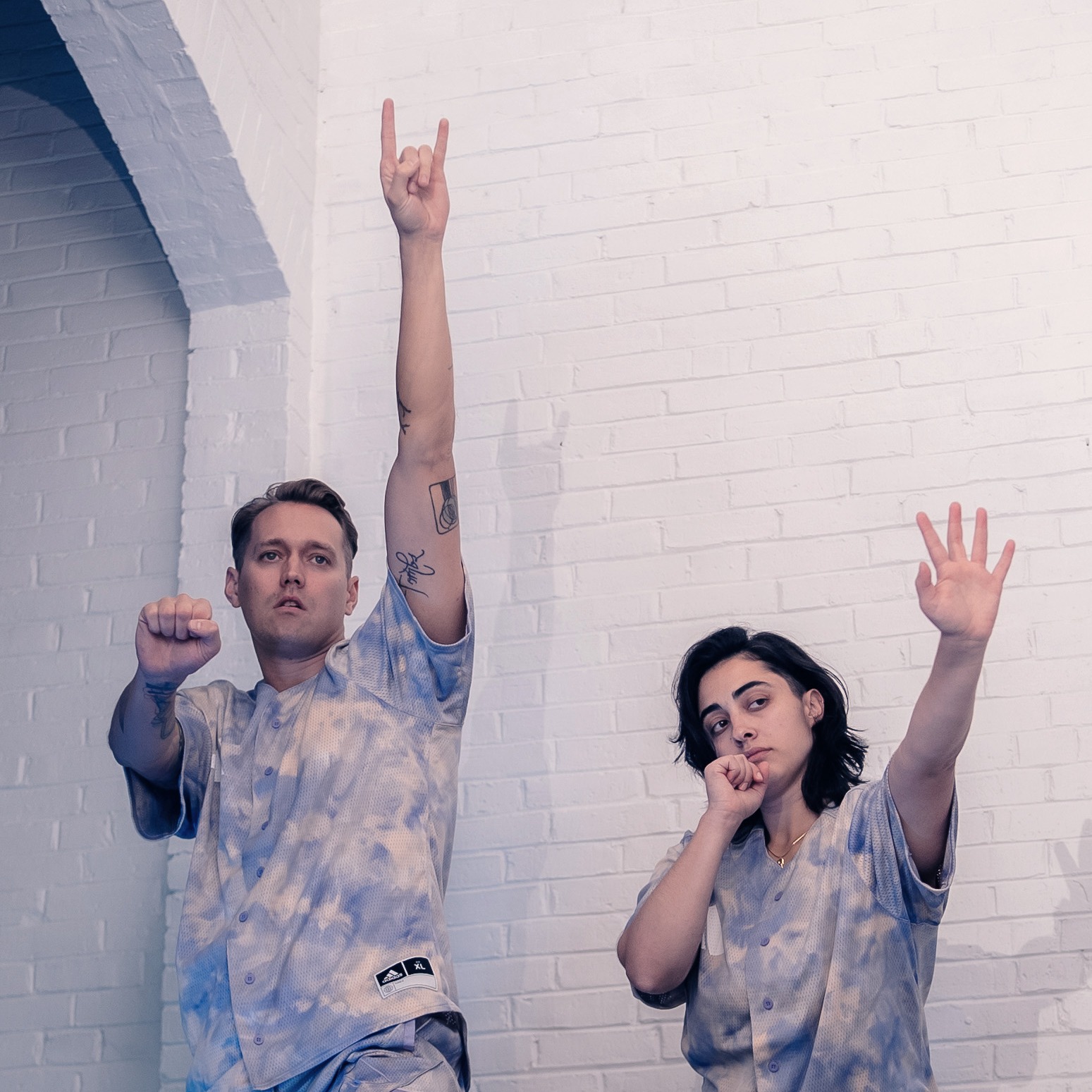 Two humans, in hazy purple baseball jerseys, stand against white brick. They raise their arms gesturing like rock stars, with a hazy and syrupy gaze.