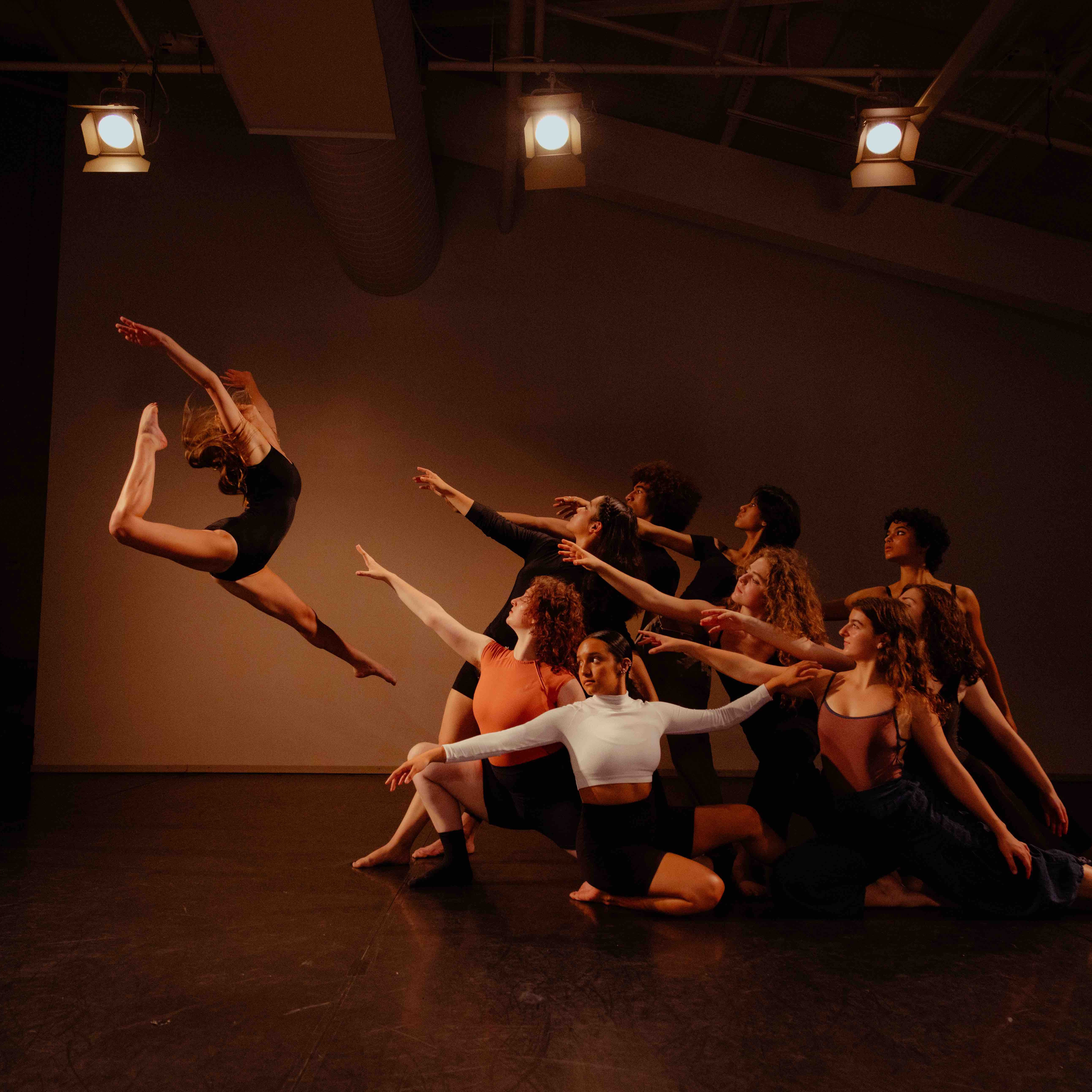 A dancer performs a leaping action while a group of dancers pose on the floor reaching towards her.