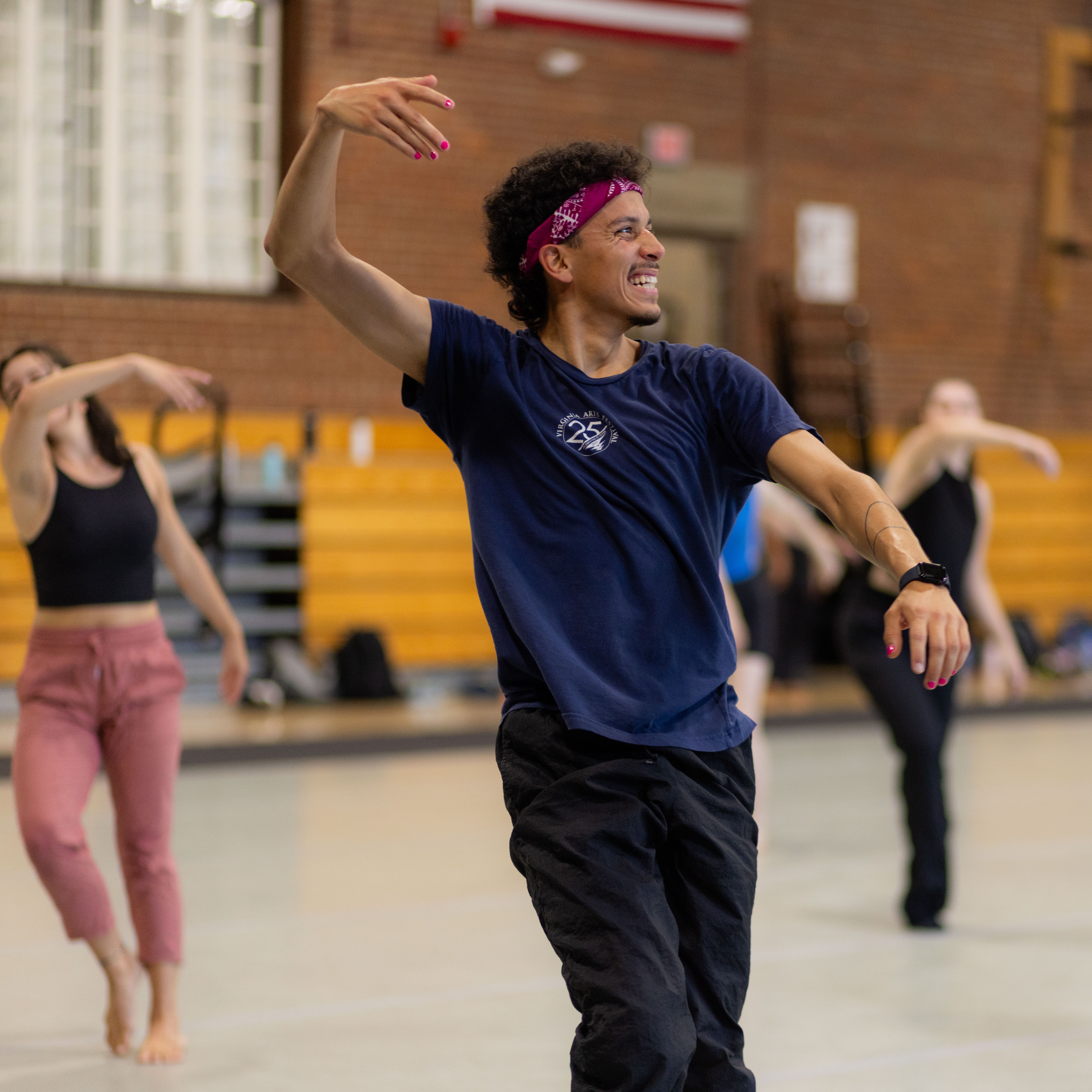 A photo of a person teaching a dance class in a gymnasium. They smile while looking away from the camera and at the students behind them.