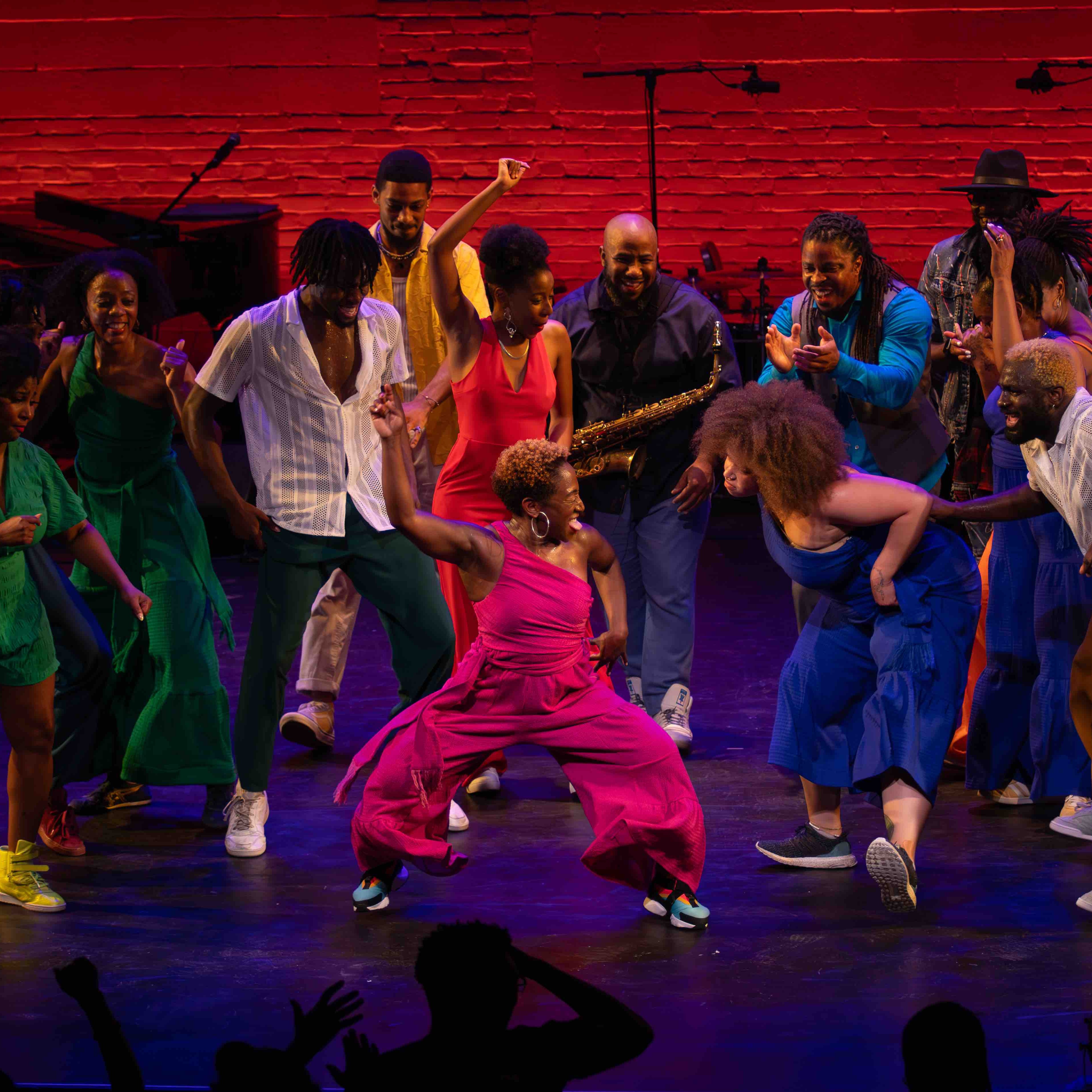 A large group of people dance, smile, and celebrate together on a colorfully lit stage.