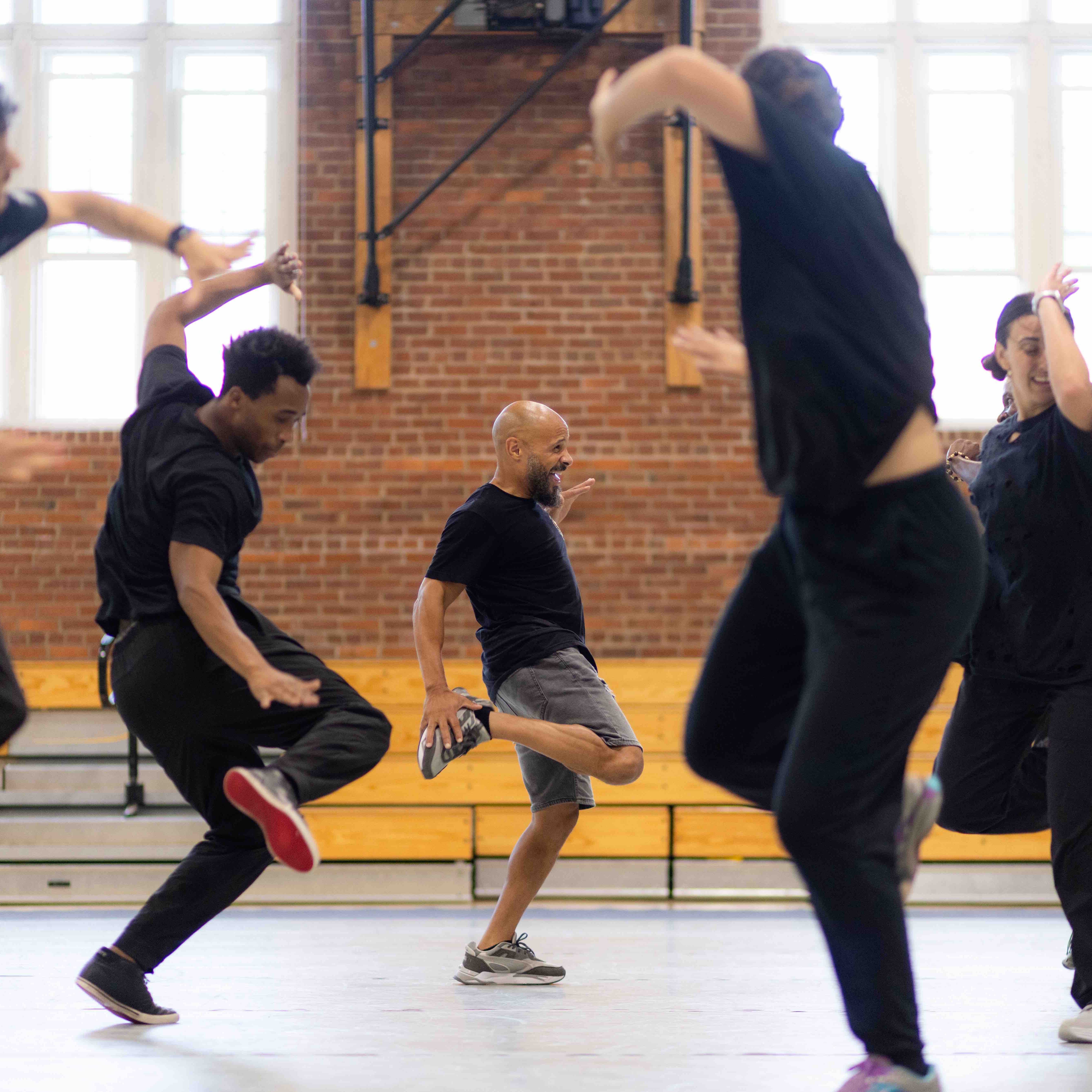 A group of people wearing all black perform a dance in a brightly lit gymnasium space.