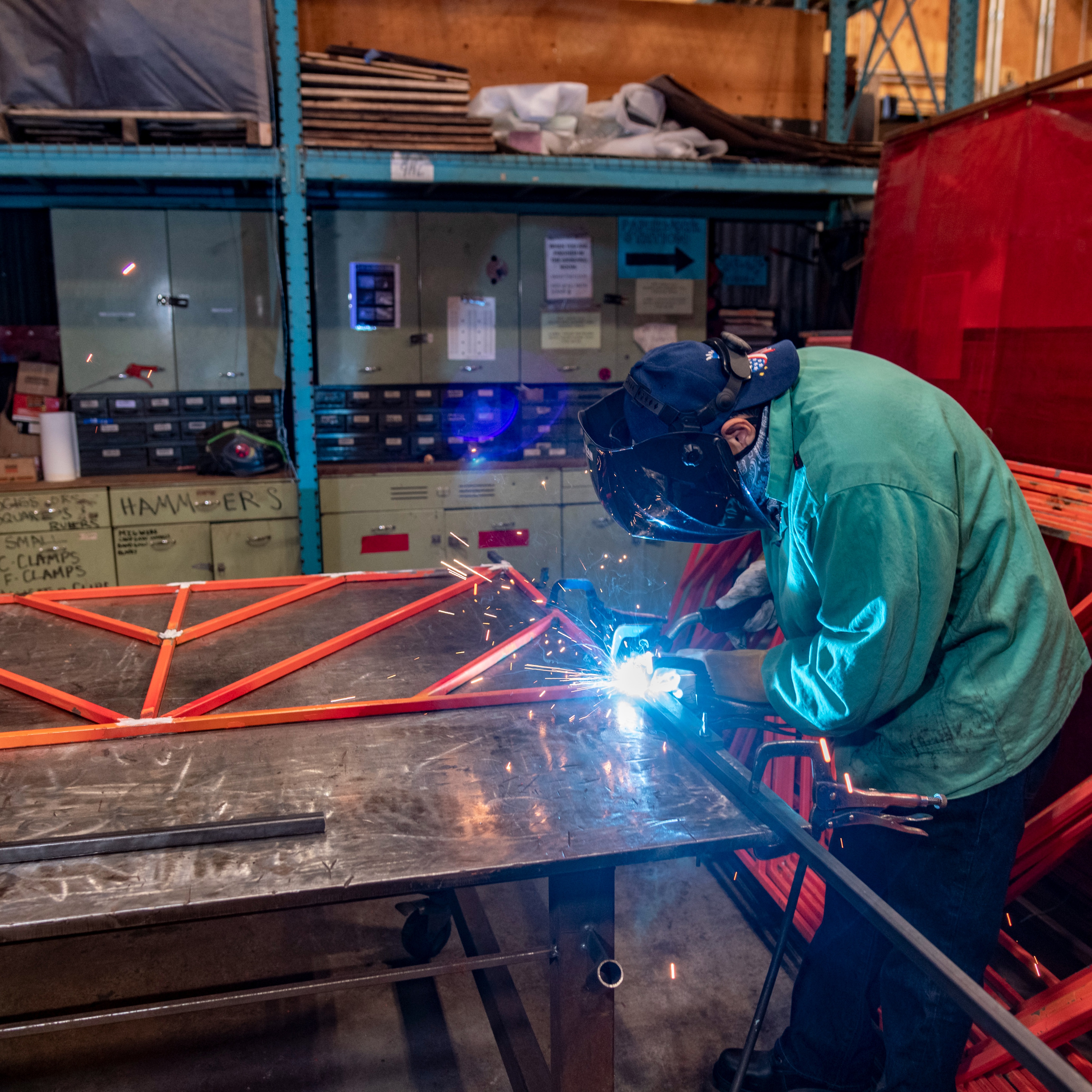 Public Projects Apprentice welds connective pieces for a bright orange bike rack.