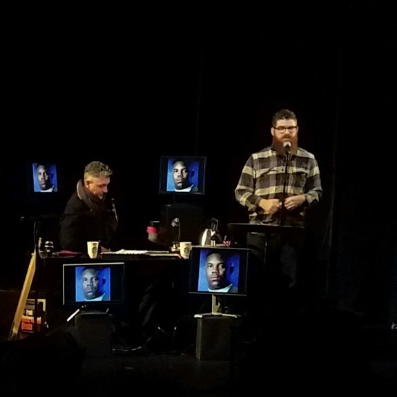 The stage is black and three male figures are illuminated along with approximately a dozen television monitors displaying the same headshot of a black man.  