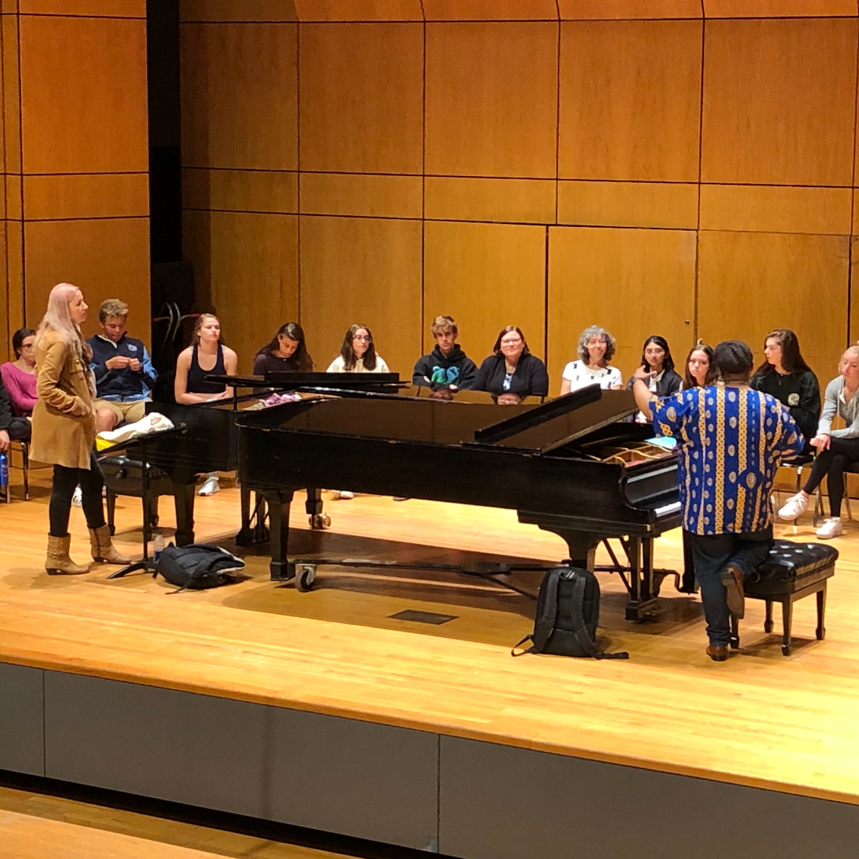There are two pianos center stage, one pianists a tall white woman with blond hair stands on the left of the pianos and the another pianist an black African man stands to the right of the pianos.  The is group of students seated in an arch upstage. 