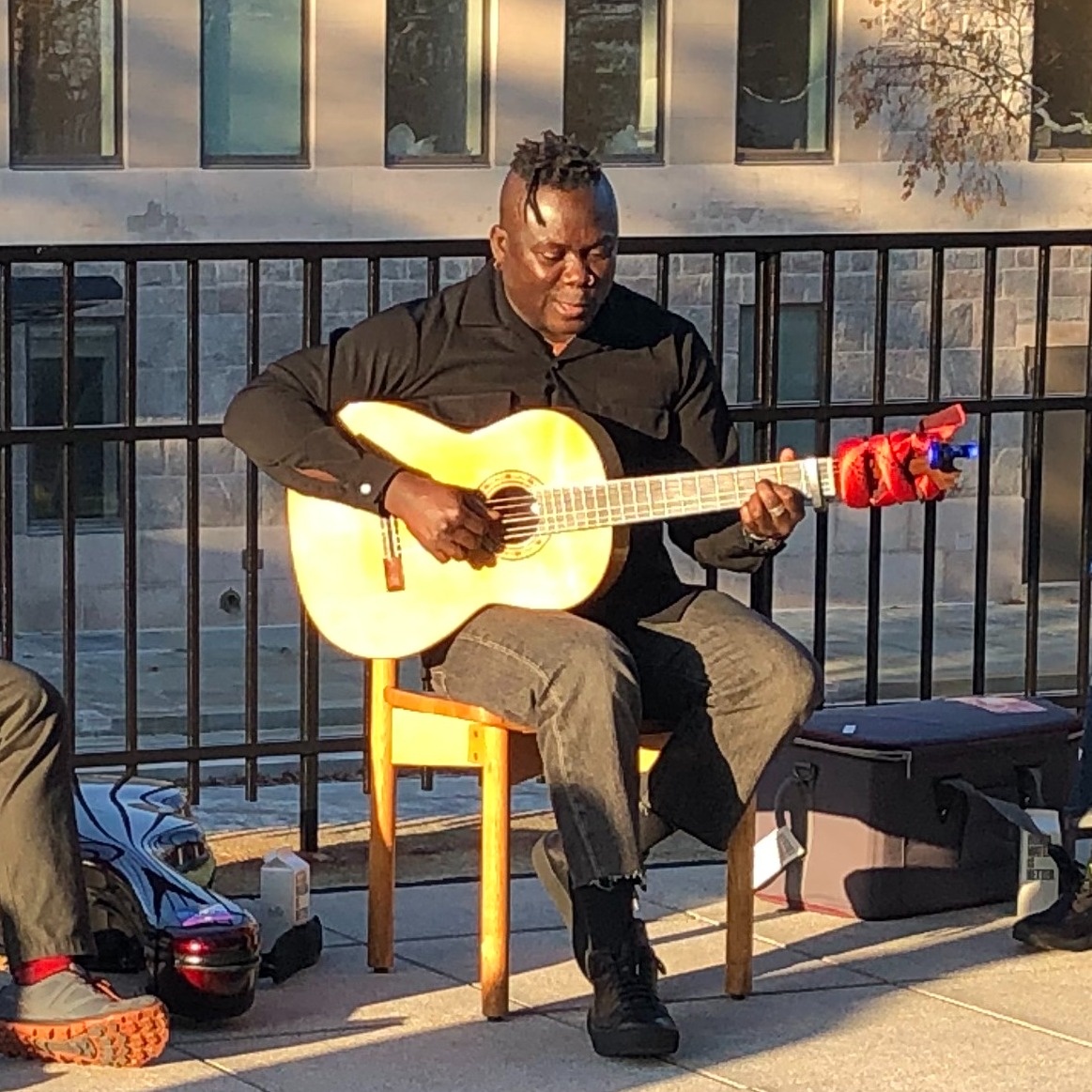 Ghanaian musician seated outside playing his guitar with a percussionist on either side with students in the foreground.