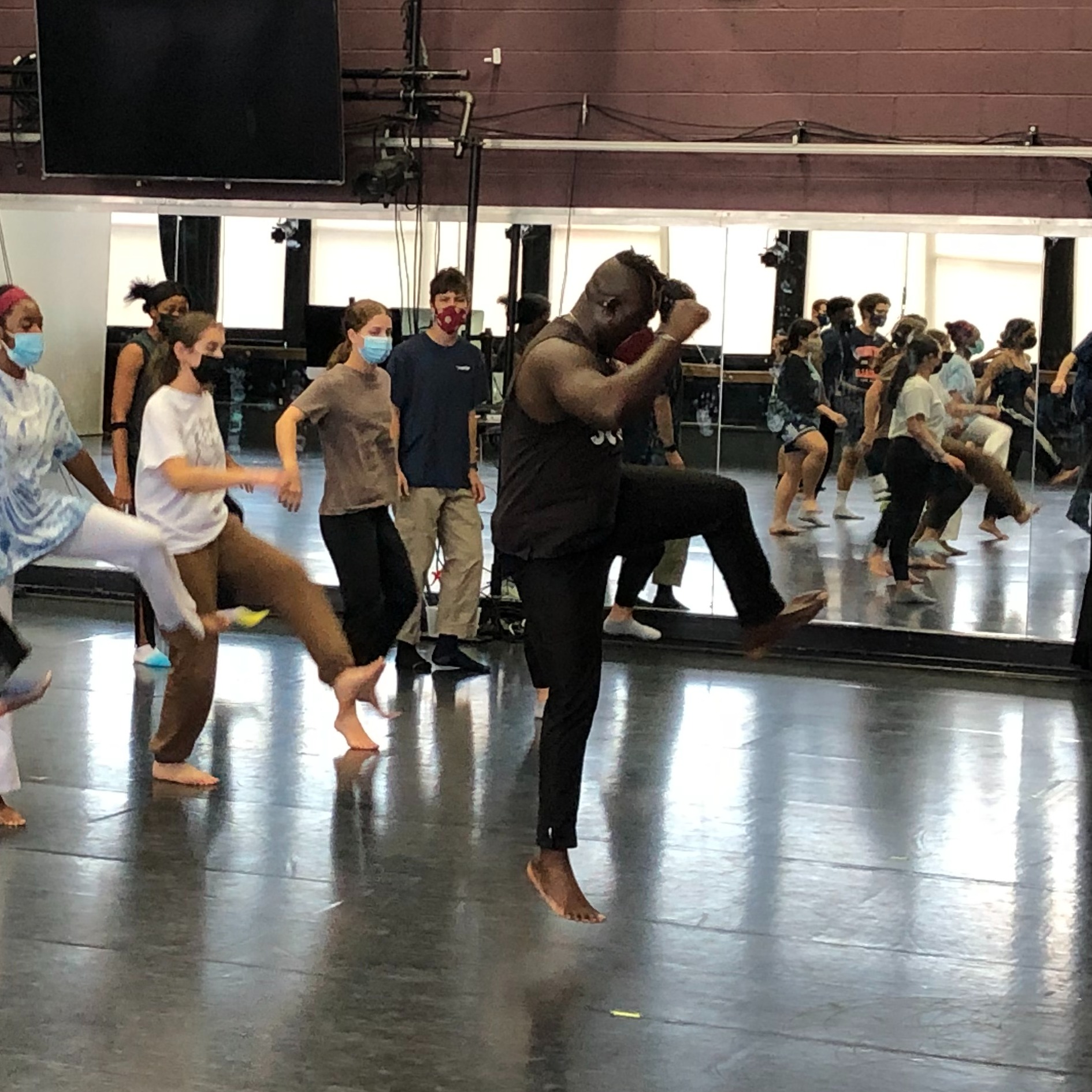 Ghanaian dancer leaping in the air with one knee raised as he leads dance class with a group of students behind him.