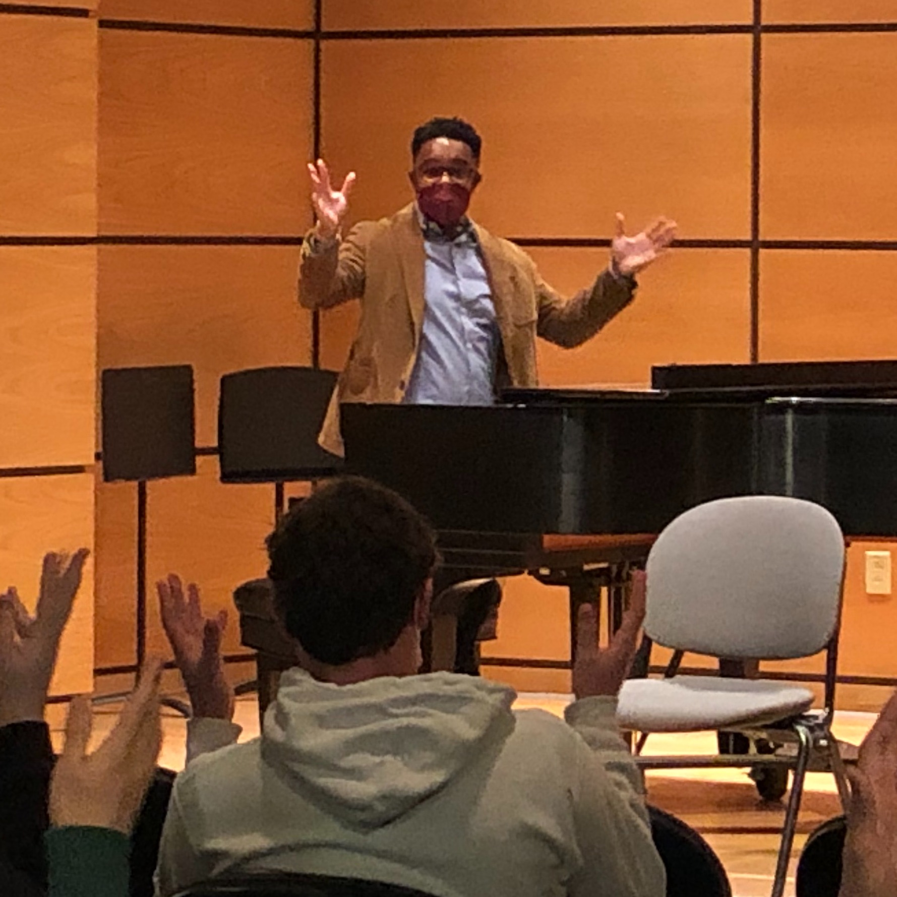 African American musician standing at a piano while he teaches a class for students in the foreground.