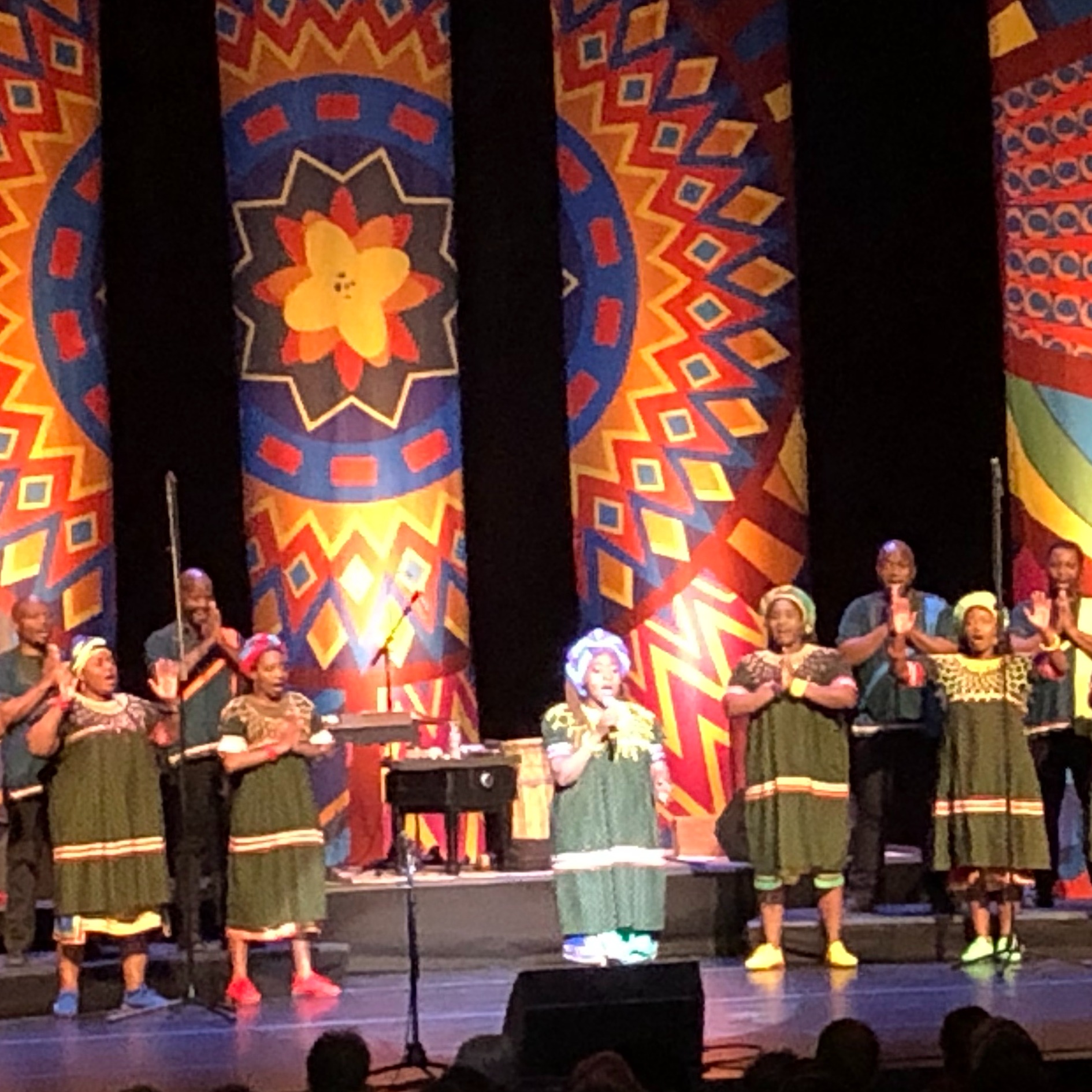 The South African Soweto Gospel Choir standing on stage during a concert with bright colored backdrops. 
