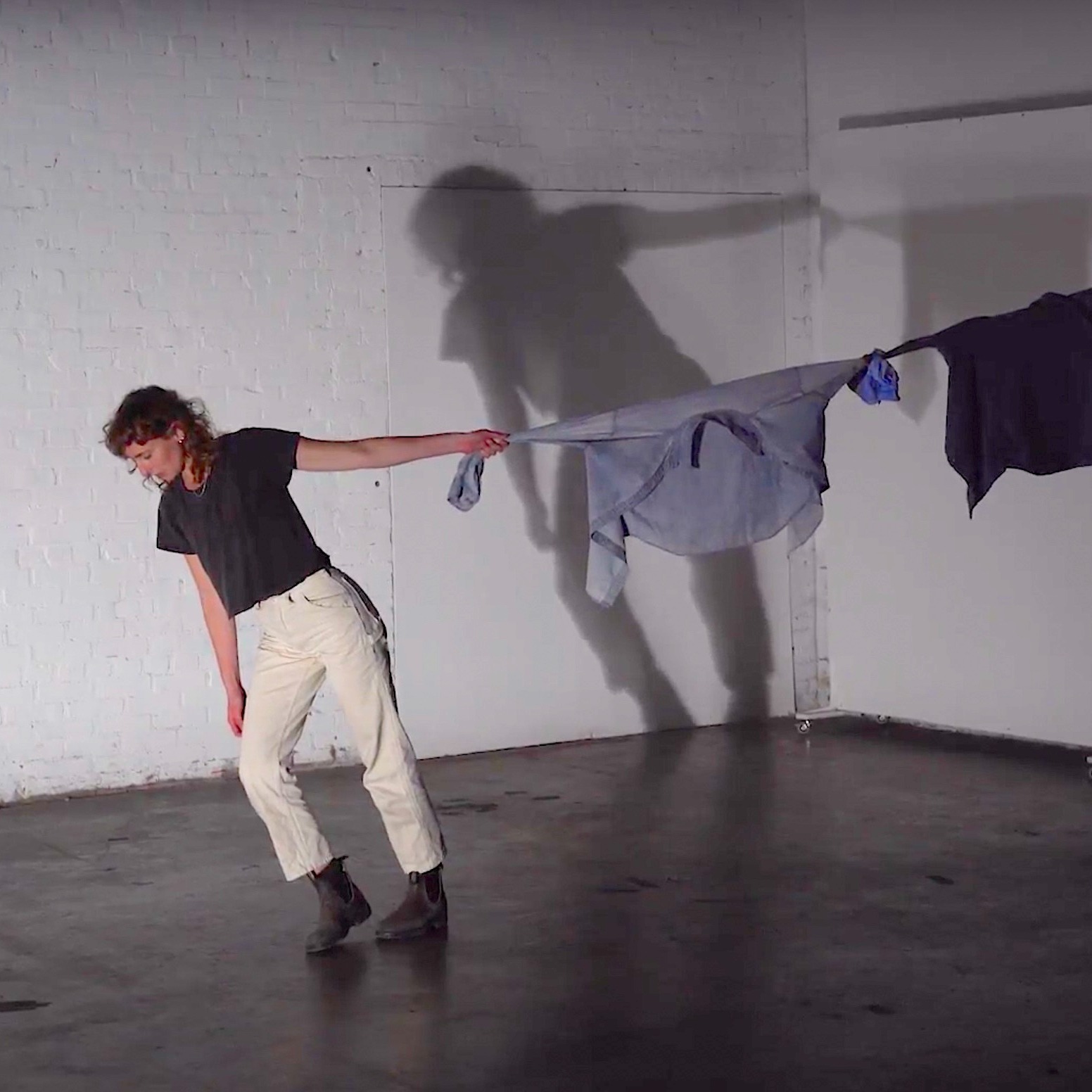 A white dancer with light brown curly mid-length hair dressed in white pants, black tee shirt and black boots holds onto a string of shirts tied to a beam in the space. She leans away, looking down at the ground and pulling the shirts taut.