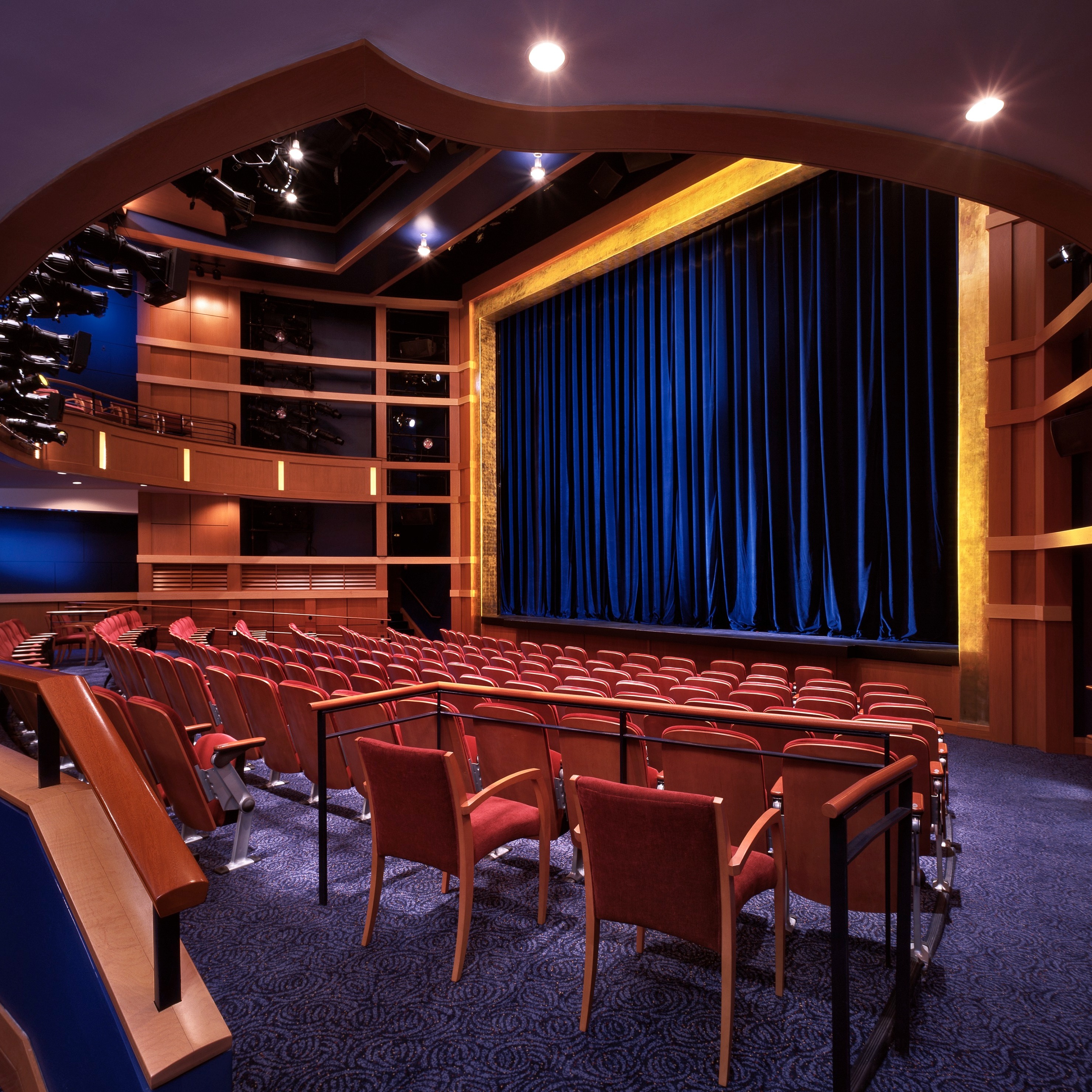 An intimate theater space with blue velvet curtain, gold leaf proscenium, warm wood walls, red seats and blue patterned carpet.